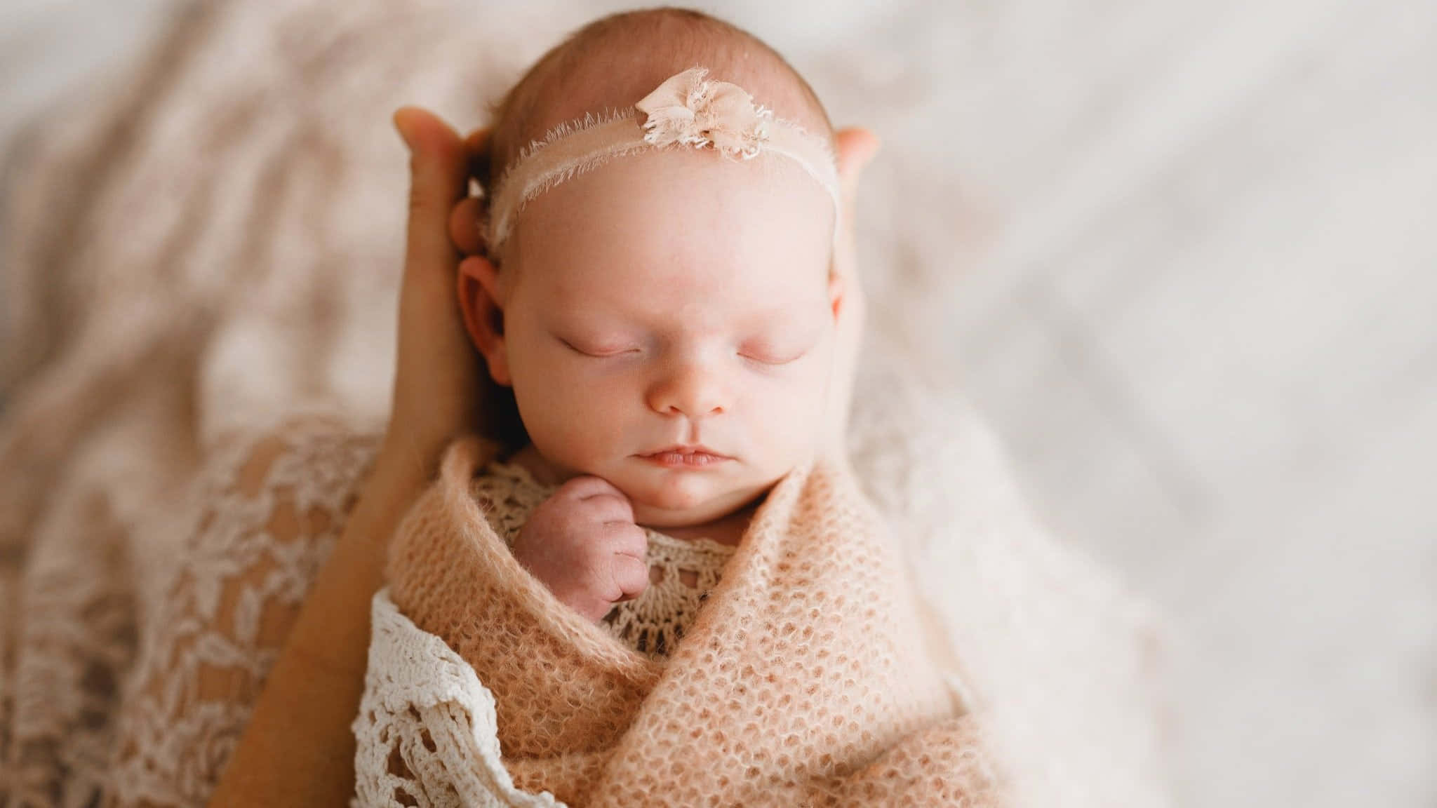 Newborn Baby 桌布 壁紙 — 一個寧靜的新生嬰兒被柔軟的蜜桃色針織布包裹著，配有蕾絲邊和精緻的頭帶，安詳地睡著。
