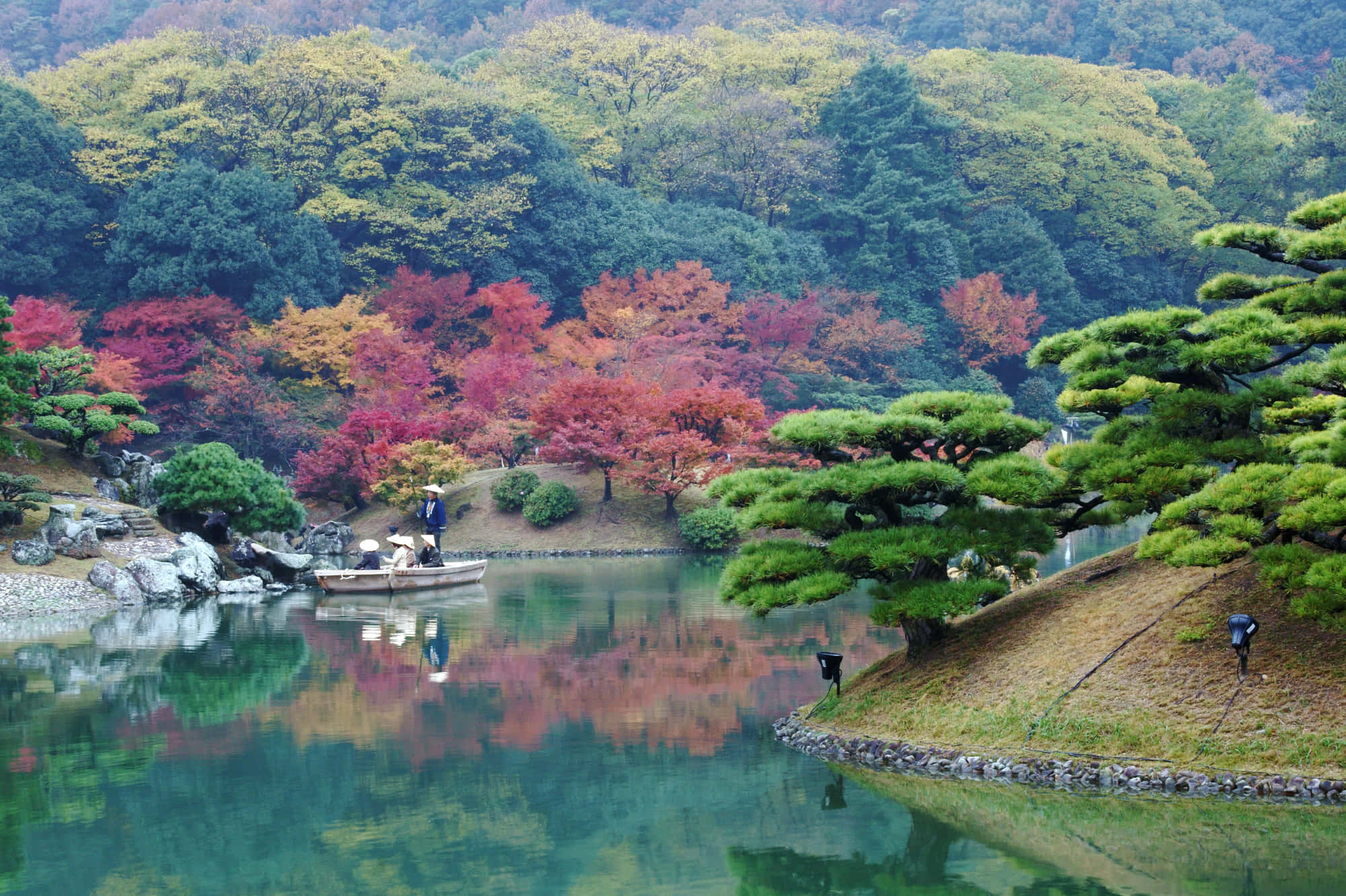 Samurai Garden 桌布 壁紙 — 一個寧靜的日本花園池塘倒映著充滿活力的秋葉，以及一艘孤獨的小船，船上有一人在多雲的天空下划船。