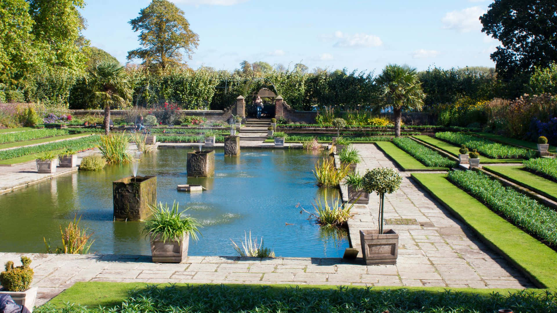 Garden Design 桌布 壁紙 — 一個寧靜的正式花園，中央有一個池塘，周圍是修剪整齊的草坪和石徑，上方是明亮的藍天。