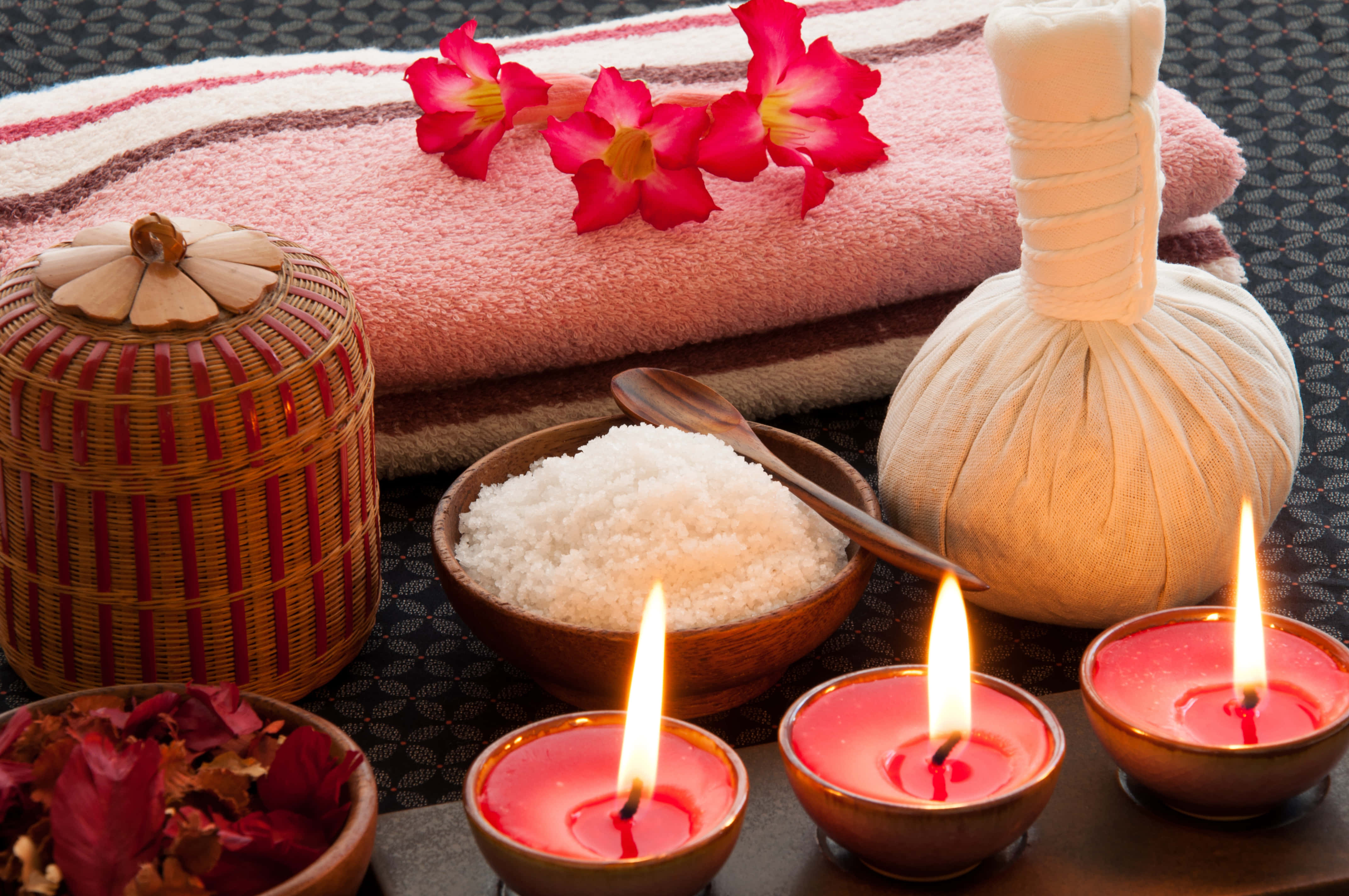 Spa Treatments 桌布 壁紙 — 一個寧靜的水療佈置，配有點燃的紅色蠟燭、鹽碗、按摩袋和毛巾上的粉紅色花朵。