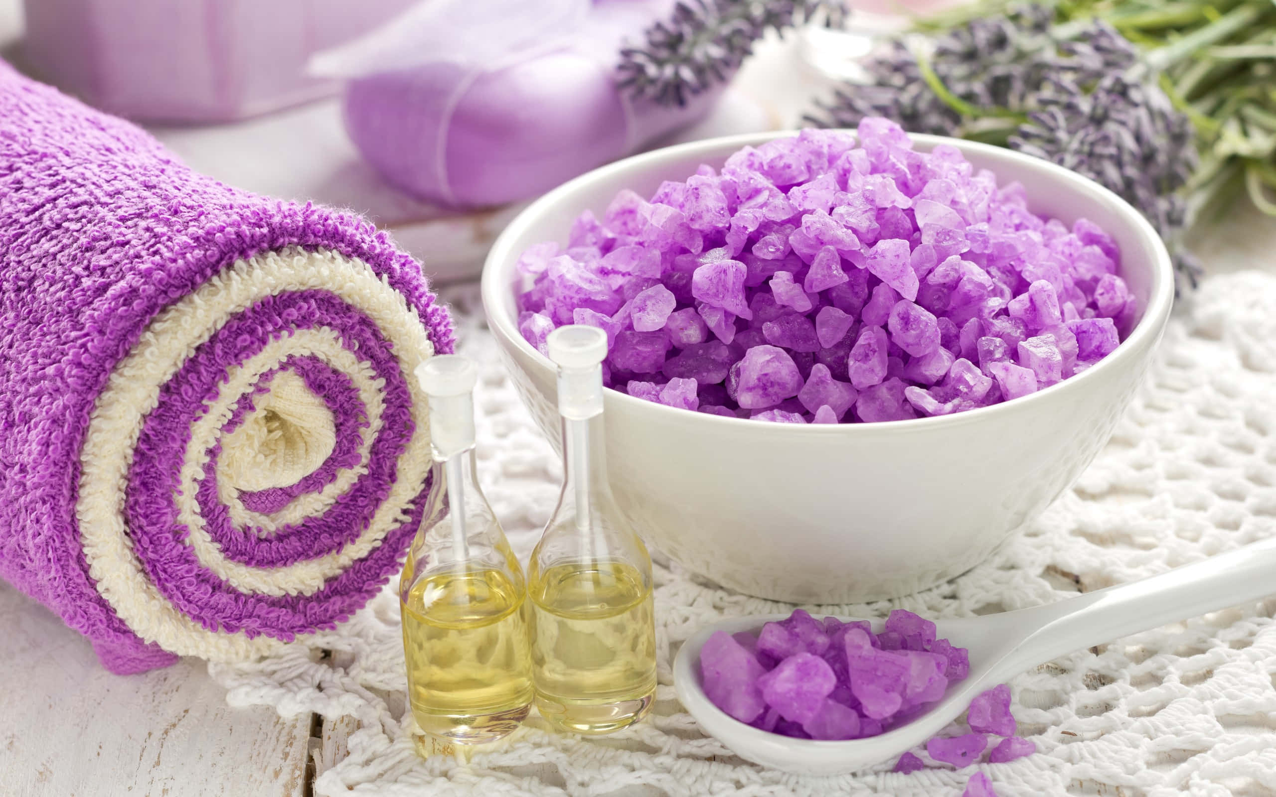 Spa Treatments 桌布 壁紙 — 一個寧靜的水療設置，包括紫色浴鹽、薰衣草枝、捲起的毛巾和精油，放在白色蕾絲表面上。