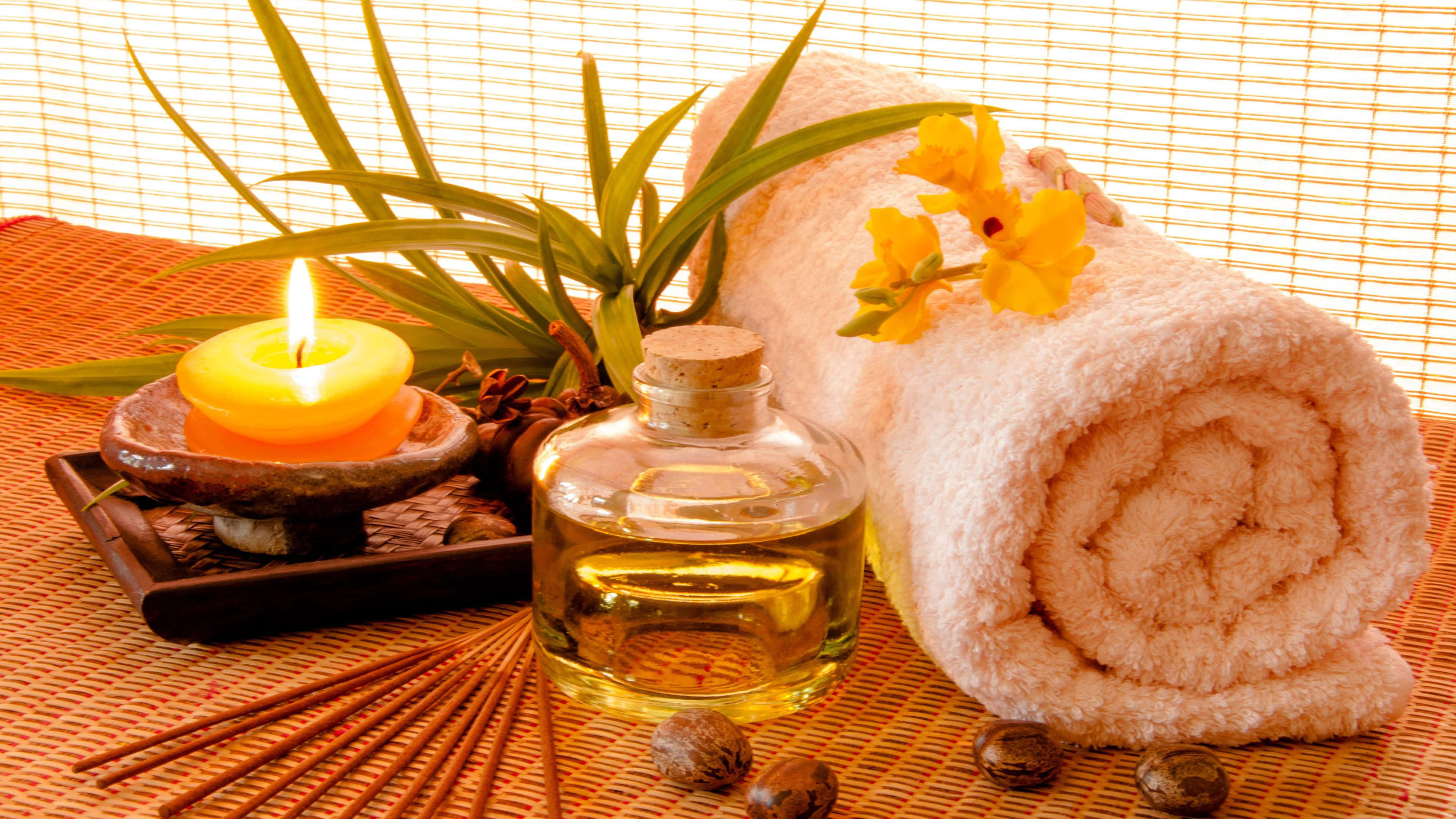 Spa Treatments 桌布 壁紙 — 一個寧靜的水療設置，包括點燃的蠟燭、捲起的毛巾和精油瓶，放在編織墊上。