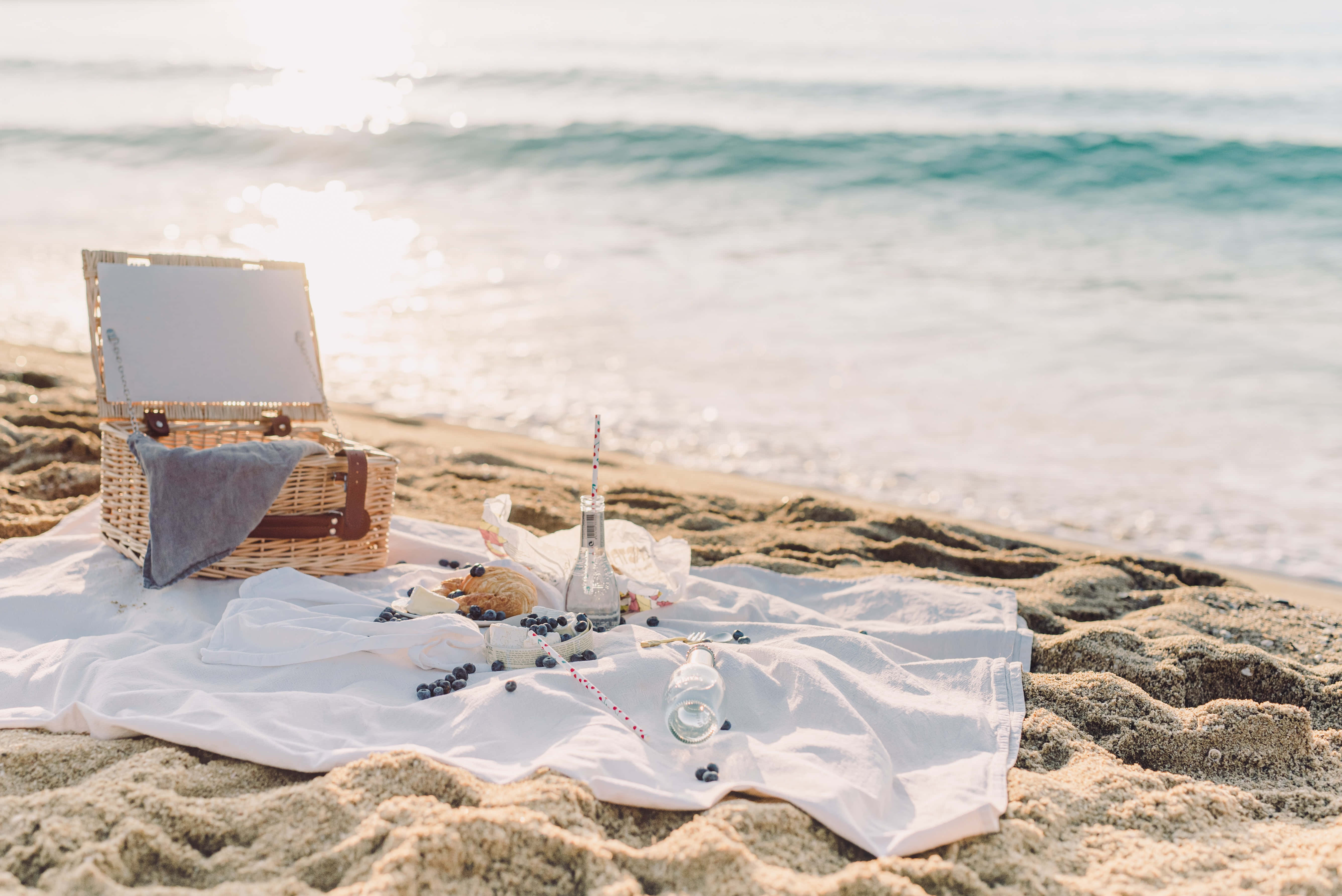Beach Picnic 桌布 壁紙 — 一個寧靜的海灘野餐佈置，在白色布上放著柳條籃子和藍莓，溫柔的海浪在黃金時段緩緩湧來。