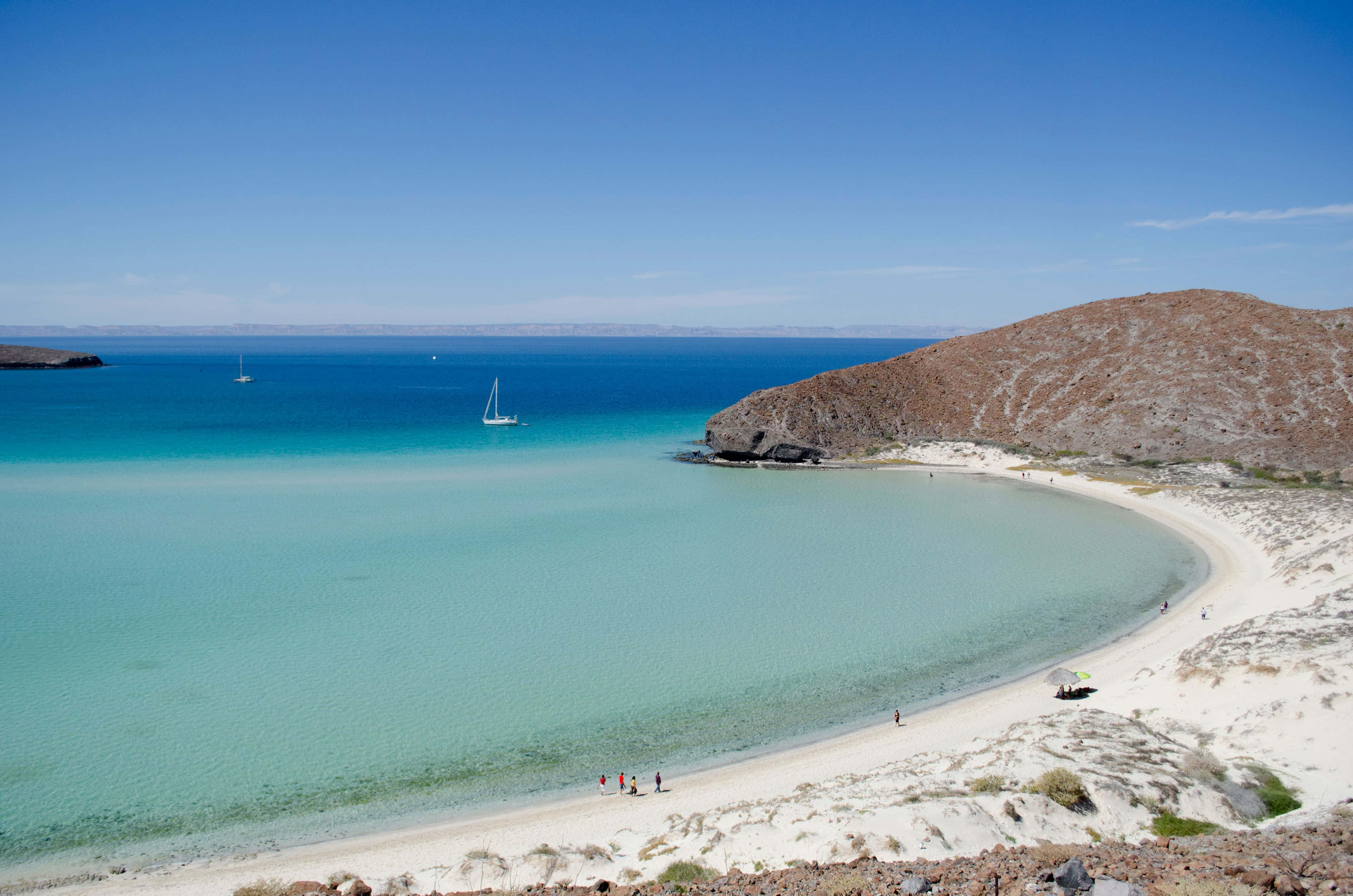 Mexican Beach 桌布 壁紙 — 一個寧靜的海灣，碧綠的海水環繞著白色沙灘，兩旁是乾旱的山丘，上方是清澈的藍天。