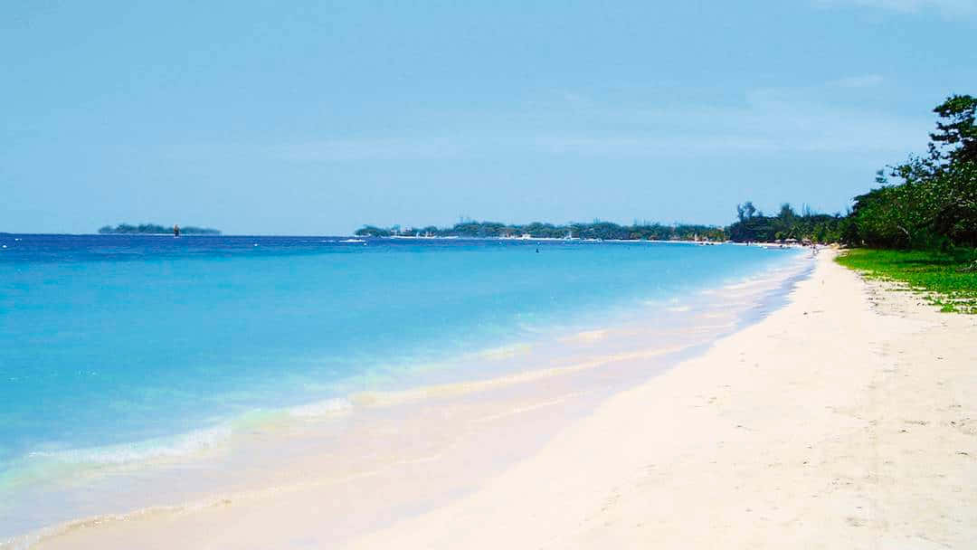 Jamaican Island 桌布 壁紙 — 一個寧靜的熱帶海灘，擁有粉狀白色沙灘和清澈碧綠的海水，在明亮的藍天下。