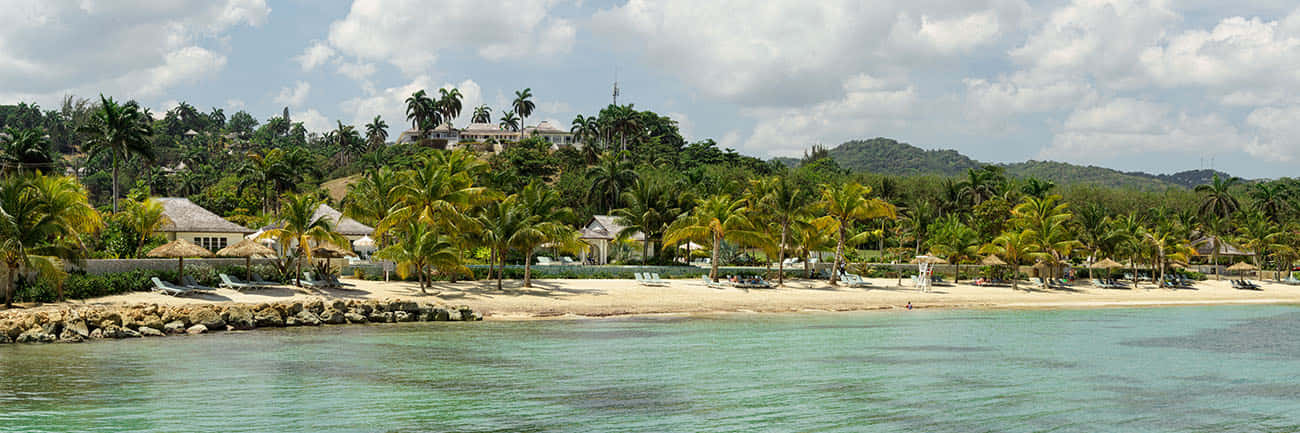 Jamaican Beach 桌布 壁紙 — 一個寧靜的熱帶海灘，有碧綠的海水、棕櫚樹和度假村建築，坐落在部分多雲的天空下的沙灘上。