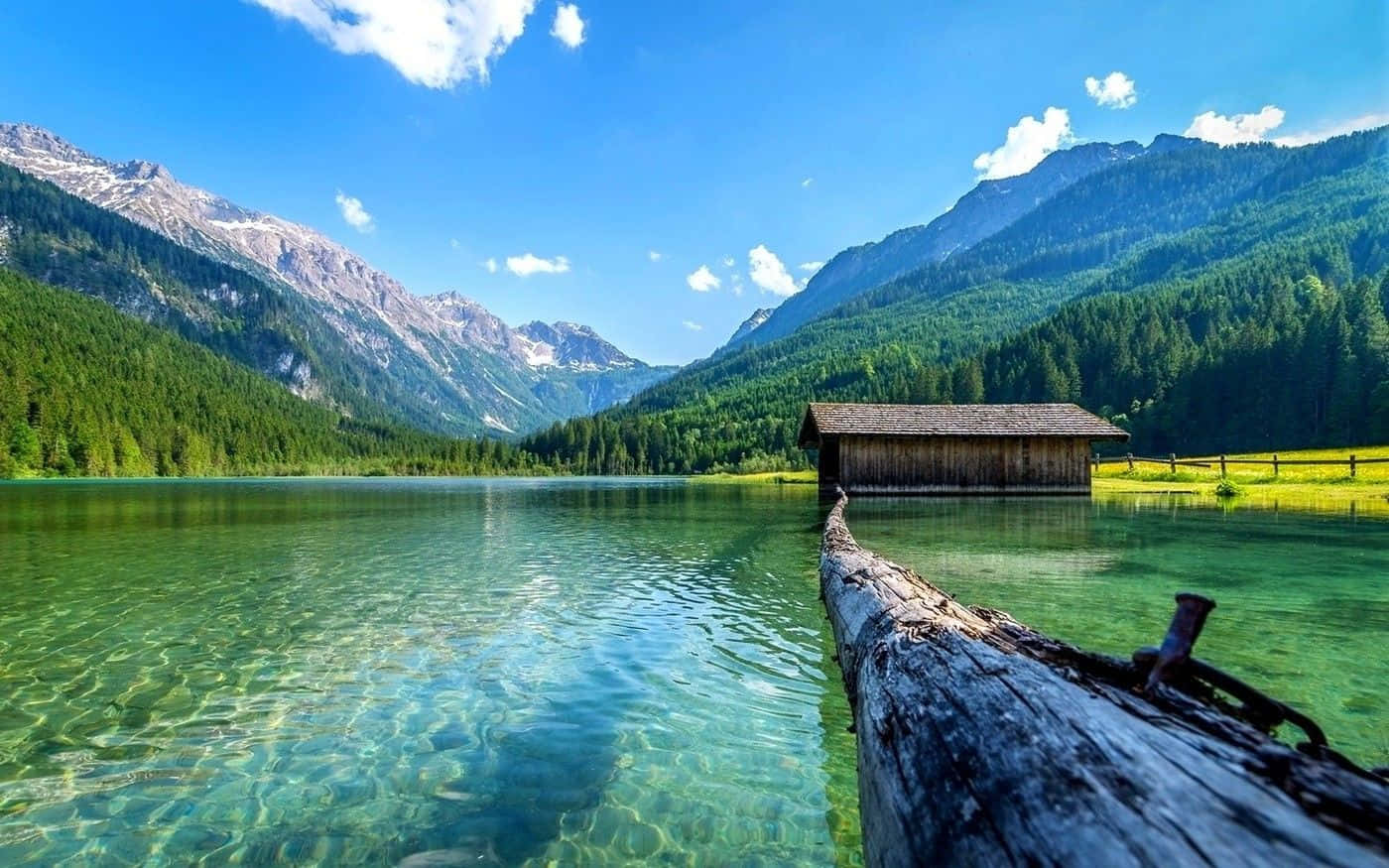 Summer Landscape 桌布 壁紙 — 一個寧靜的高山湖泊，清澈見底的碧綠湖水倒映著白雪覆蓋的山峰和一座木屋，在明亮的藍天下。