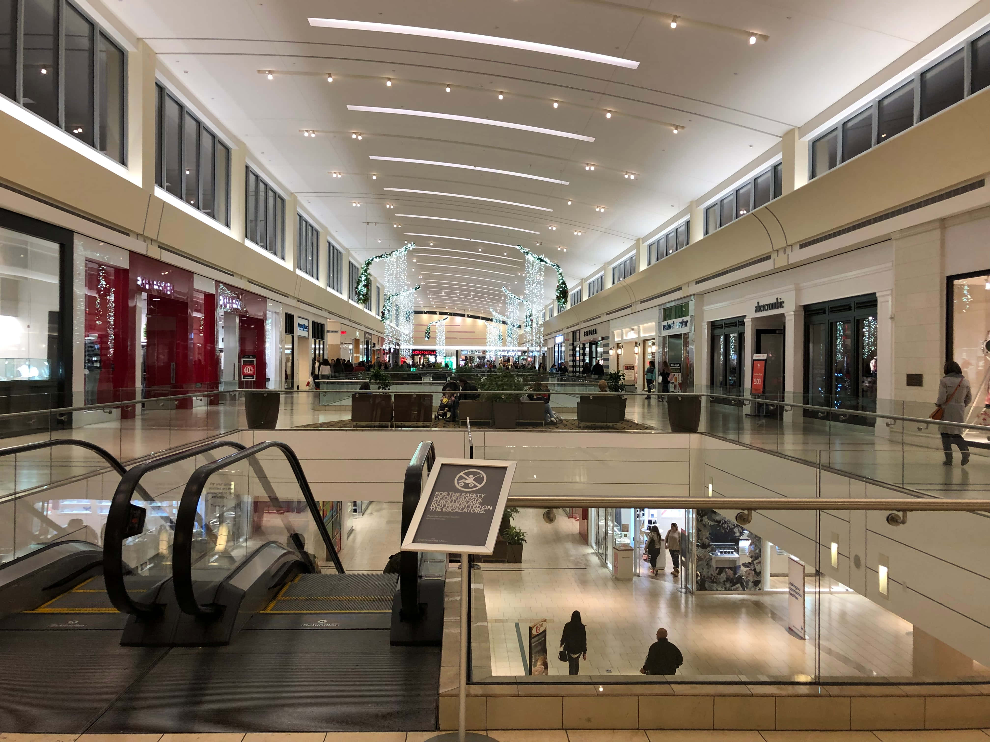 Mall 桌布 壁紙 — 一個寬敞的多層購物中心，設有自動扶梯、玻璃欄杆，購物者在明亮的吊燈下行走。
