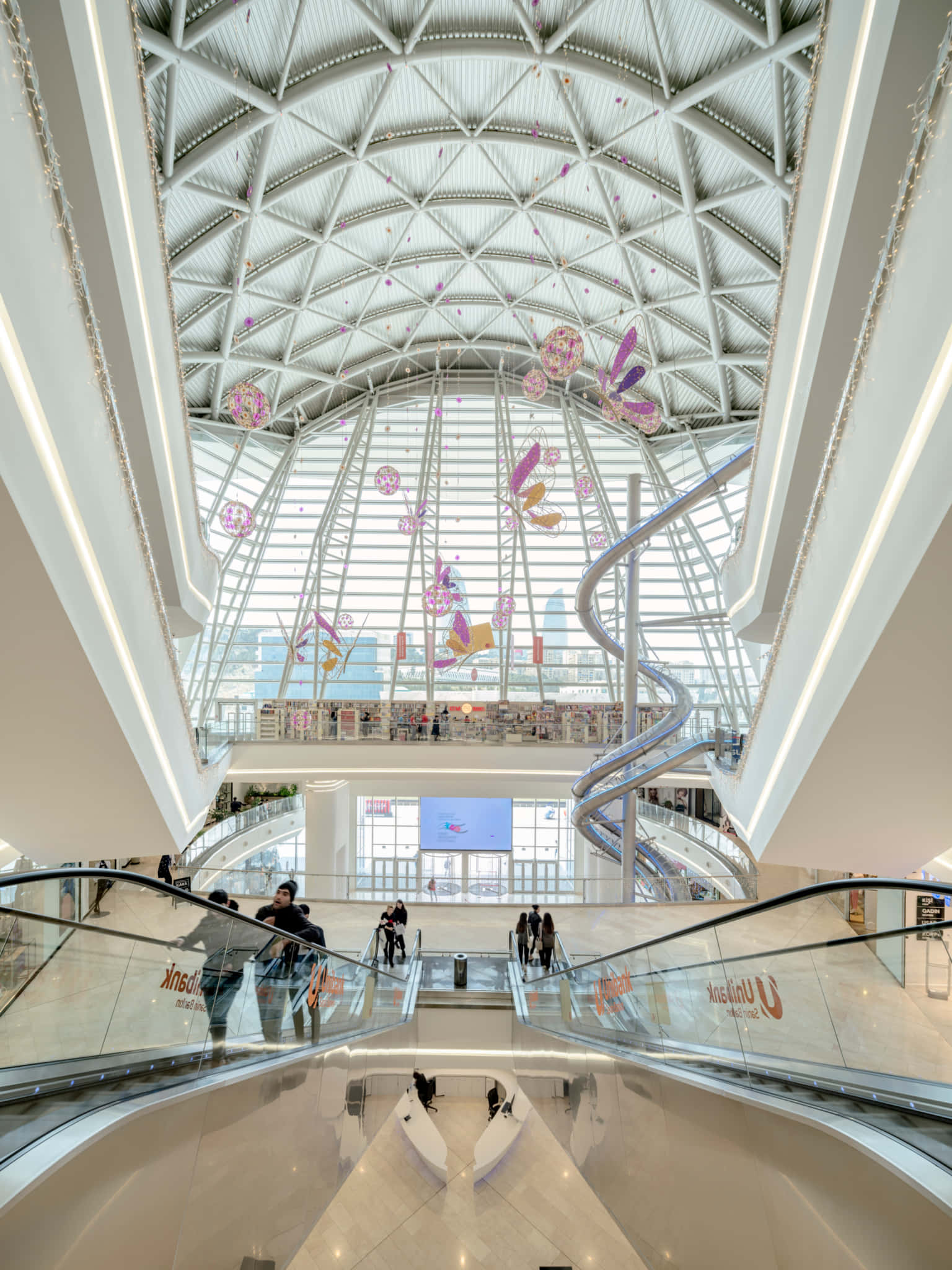 Mall 桌布 壁紙 — 一個寬敞的現代購物中心中庭，設有弧形自動扶梯、幾何玻璃天花板和色彩繽紛的蝴蝶裝飾。