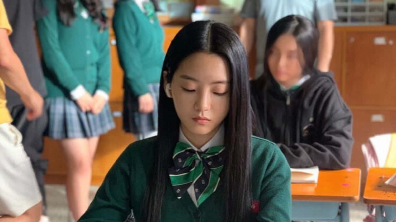 我哋都係死人 桌布 壁紙 — 一個專注的十幾歲女孩穿著綠色校服，坐在教室的書桌前，背景有同學。