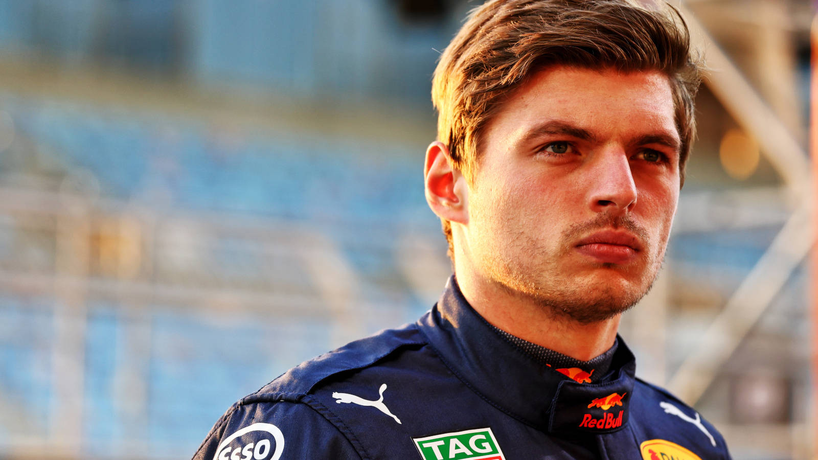 Verstappen 桌布 壁紙 — 一個專注的年輕男子穿著Red Bull賽車隊外套，在溫暖的金色光線下站在模糊的體育館看台前。