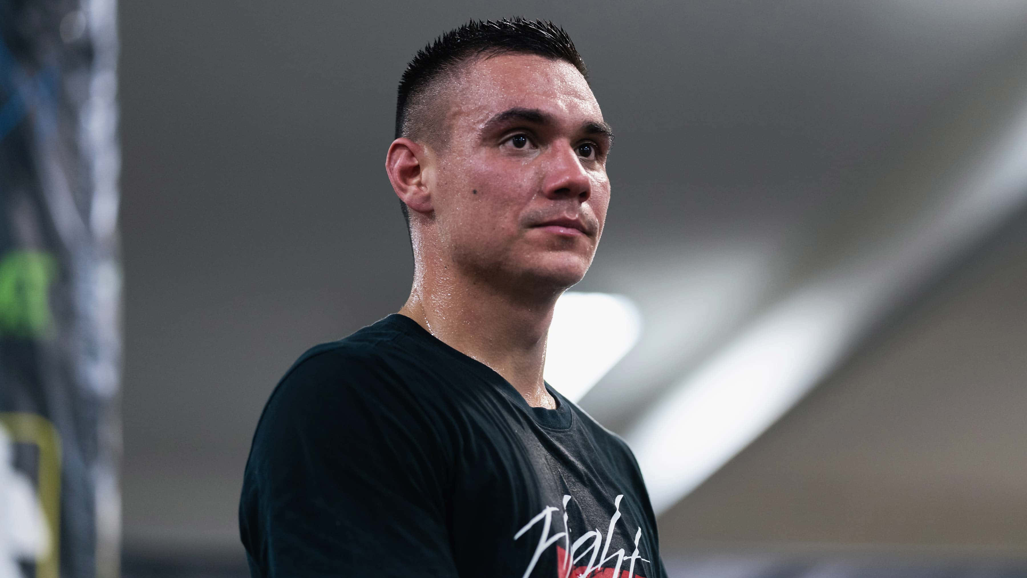 Tim Tszyu 桌布 壁紙 — 一個專注的男性戰士穿著黑色T恤，在昏暗的競技場中凝視著鏡頭外。