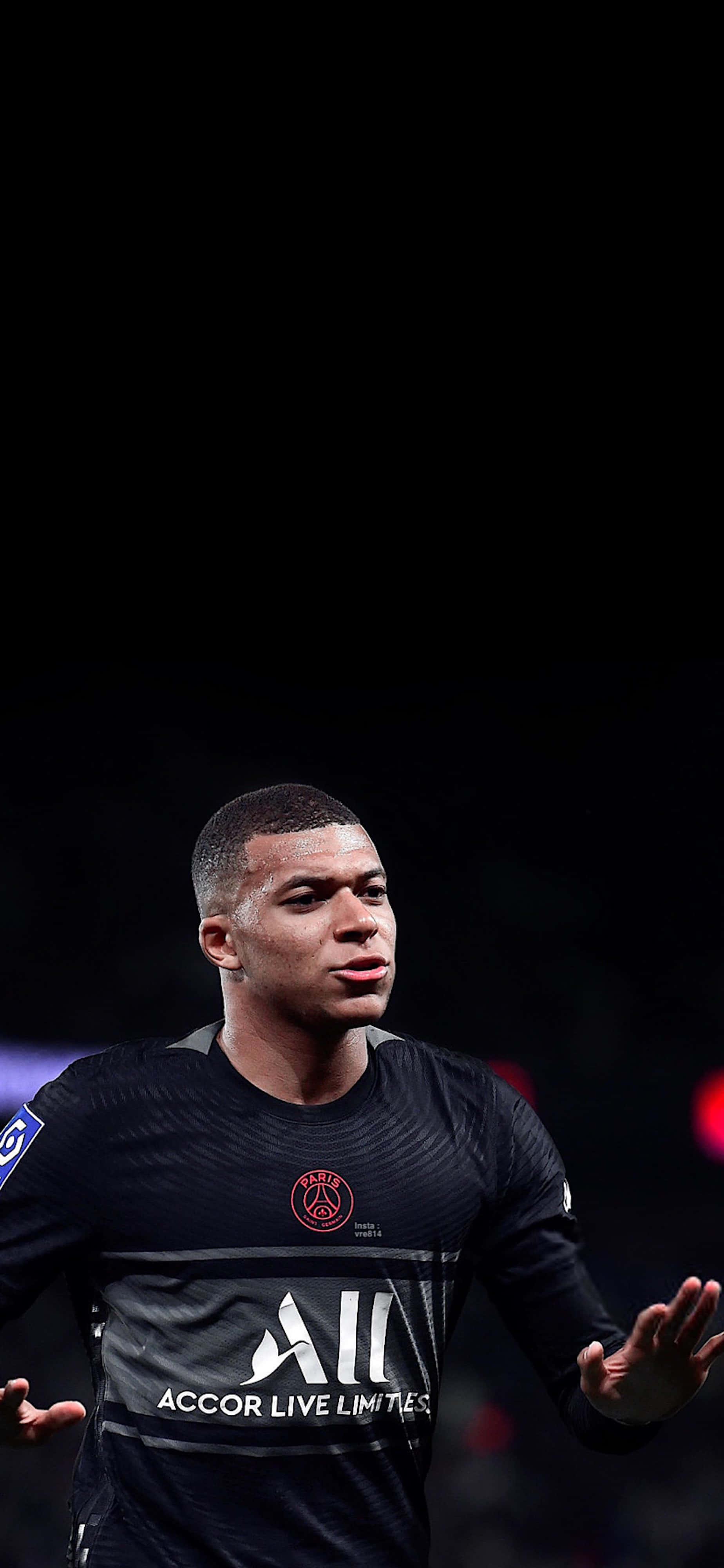 Mbappe 桌布 壁紙 — 一個專注的足球運動員穿著深色球衣，站在黑色背景前，背景有模糊的體育場燈光。