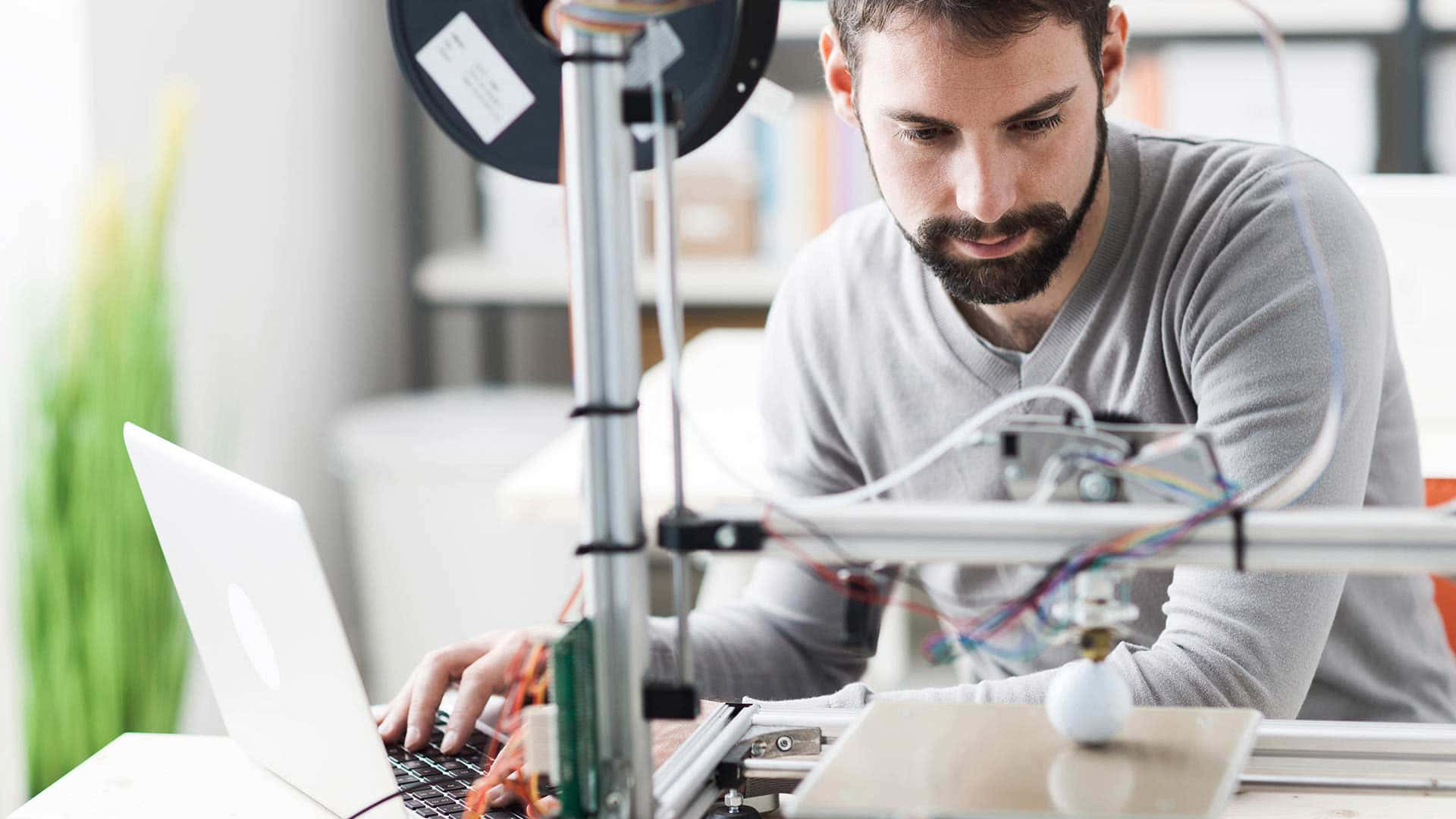 3d Printer 桌布 壁紙 — 一個專注的鬍鬚男穿著灰色襯衫，在3D打印機旁邊用筆記型電腦工作，打印機有彩色電線和一卷線材。