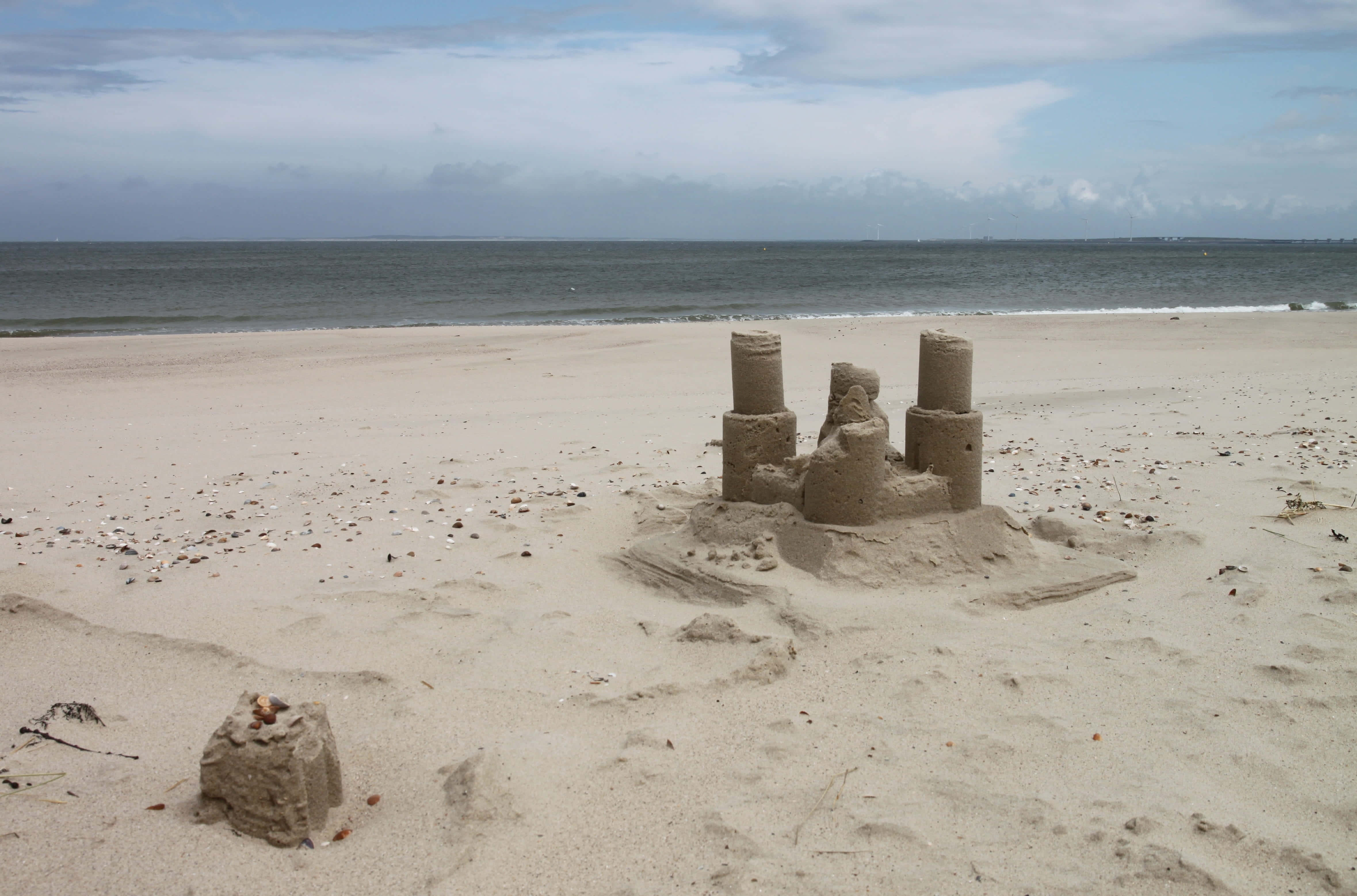 Sandcastles 桌布 壁紙 — 一個小型沙堡，有三座塔樓，矗立在寬闊的海灘上，背景是陰天和海洋波浪。