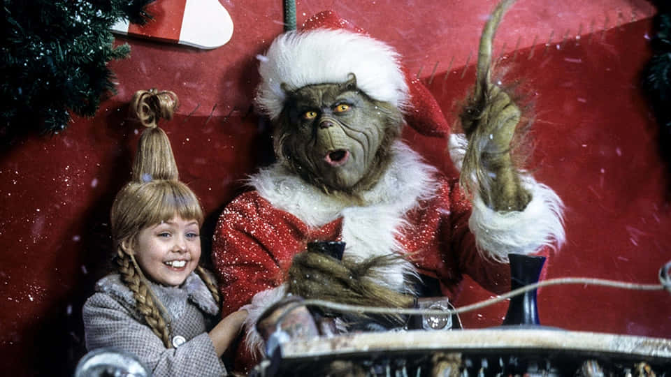 The Grinch 桌布 壁紙 — 一個小女孩在紅色雪景背景下，微笑地站在穿著聖誕老人服的聖誕怪傑旁邊。