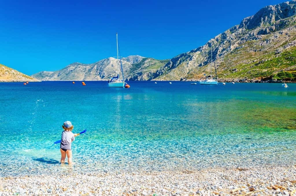 Greek Island 桌布 壁紙 — 一個小孩在卵石海灘上，於清澈碧綠的海水中嬉水，背景有帆船和崎嶇的山脈，天空湛藍明亮。