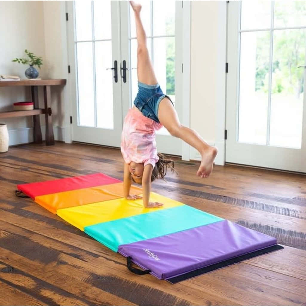 Gymnastics Mat 桌布 壁紙 — 一個小孩在陽光普照的木地板房間裡，在彩虹色的體操墊上做倒立。