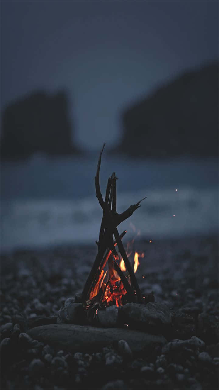 Campfire 桌布 壁紙 — 一個小營火在黃昏時分的岩石海灘上明亮地燃燒，將溫暖的光芒投射在黑暗的剪影懸崖上。
