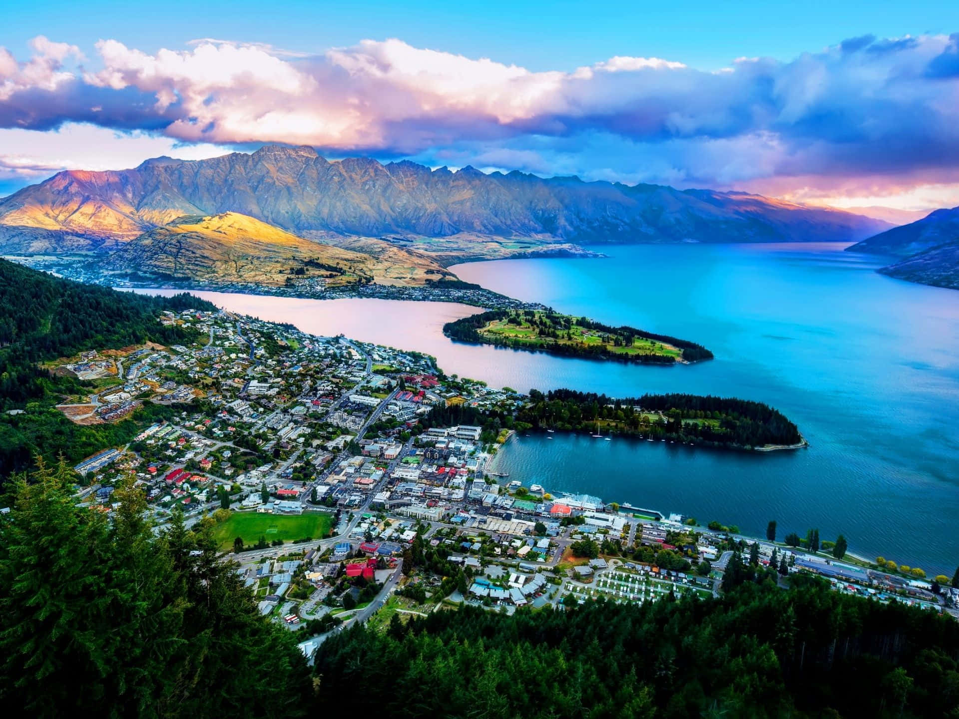 New Zealand 桌布 壁紙 — 一個小鎮的鳥瞰圖，坐落於充滿活力的藍色湖泊旁，山脈在日落色調中閃耀。