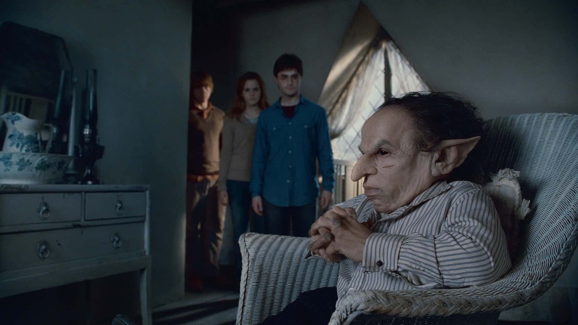 Warwick Davis 桌布 壁紙 — 一個尖耳朵嘅老妖精坐喺陰暗房間嘅藤椅上，後面門口企咗三個後生仔女。