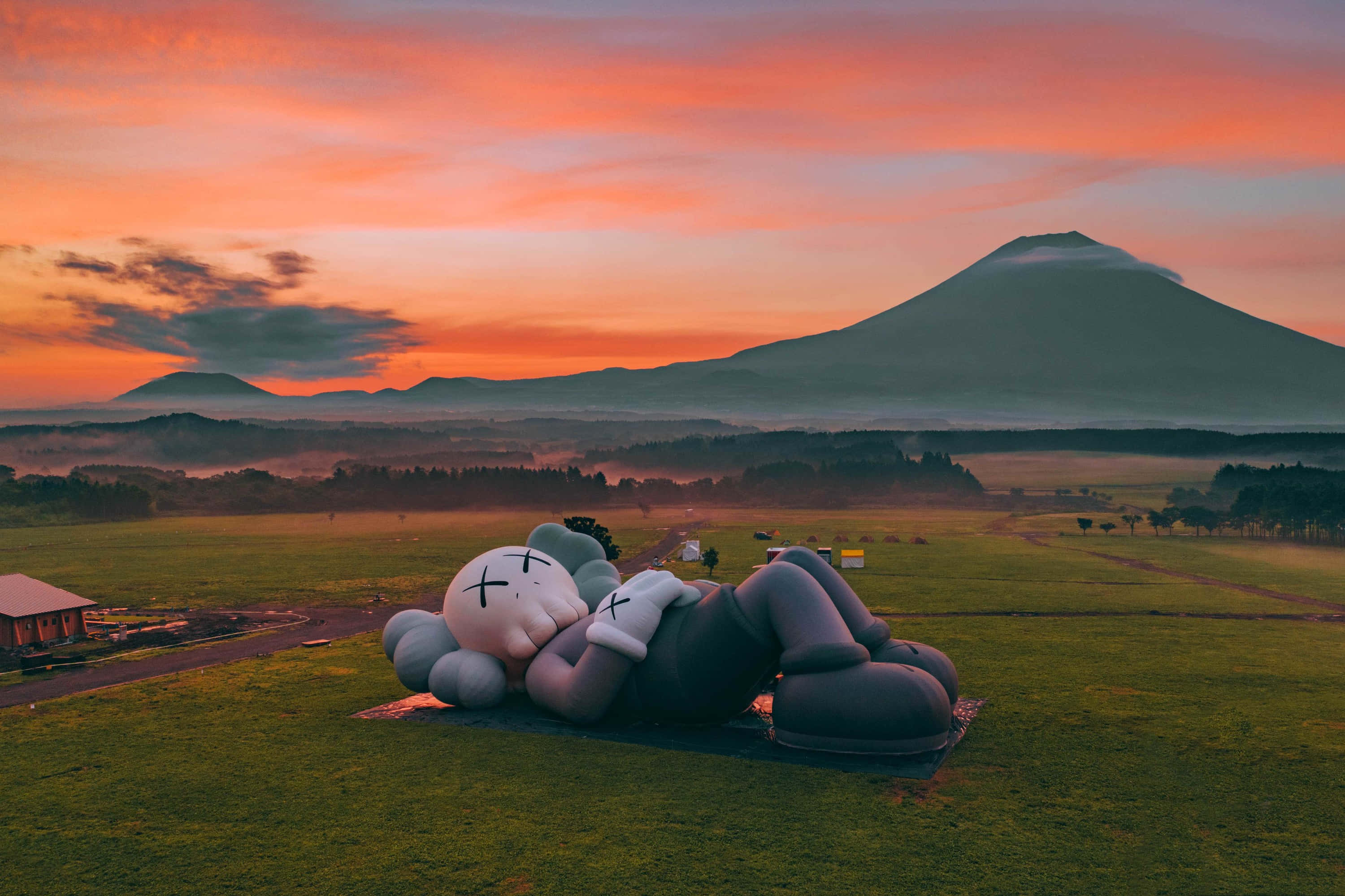 Kaws Holiday 桌布 壁紙 — 一個巨大的KAWS雕塑在日出時分平靜地躺在草地上，遠處的富士山在薄霧中若隱若現。