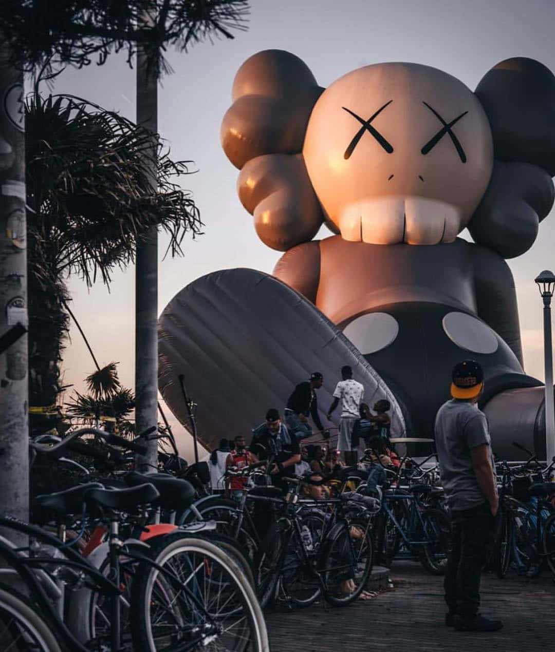 Kaws Holiday 桌布 壁紙 — 一個巨大的充氣KAWS角色在黃昏時分聳立於人群和單車之上，背景有棕櫚樹。