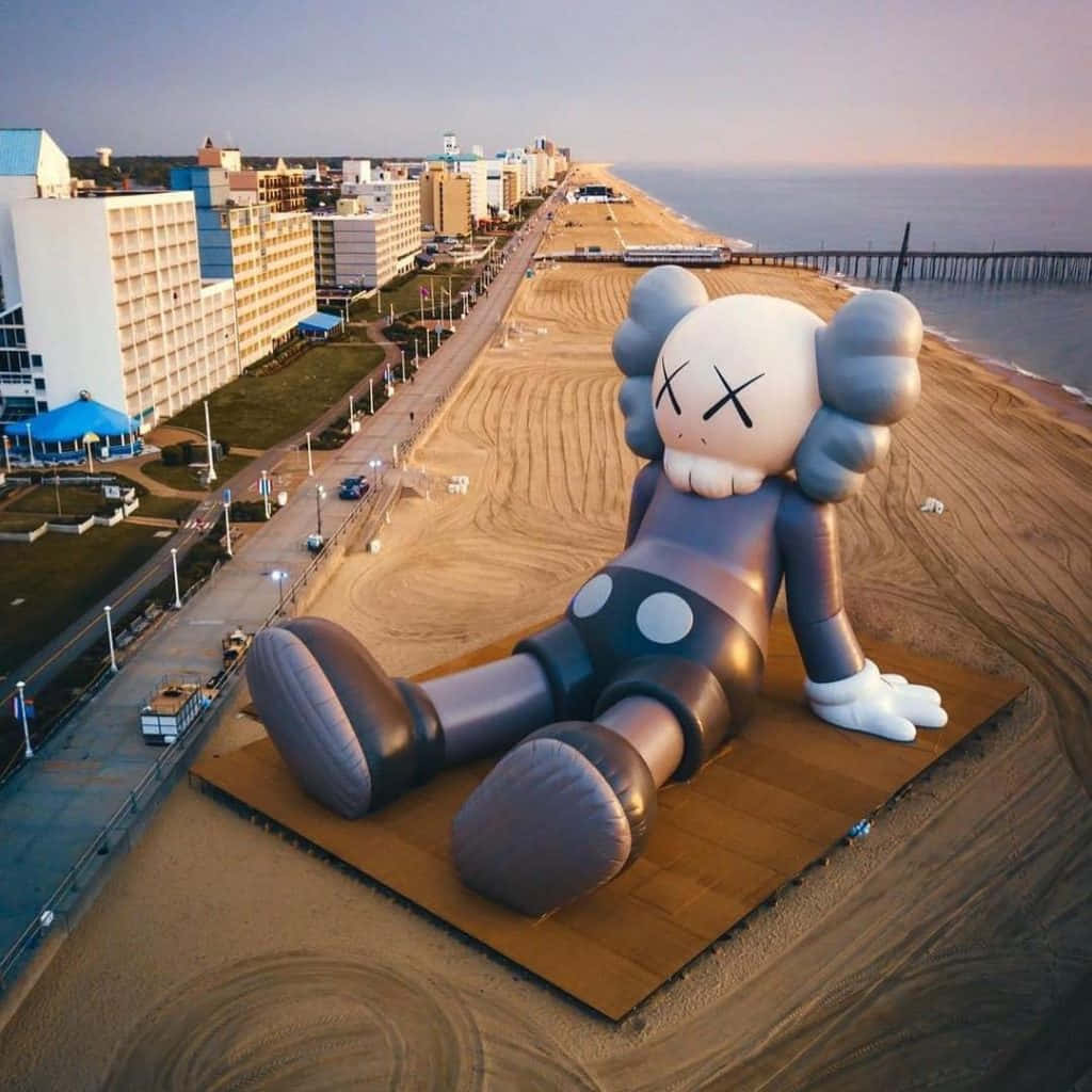Kaws Holiday 桌布 壁紙 — 一個巨大的充氣KAWS雕塑在日落時分擱在海灘上，旁邊是木板路和海洋。