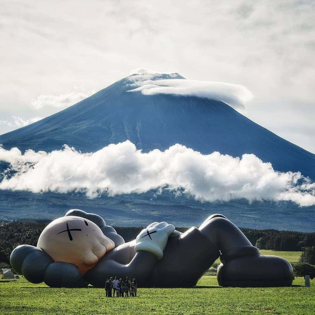 Kaws Holiday 桌布 壁紙 — 一個巨大的充氣KAWS雕塑躺在草地上，背景是富士山和雲朵。