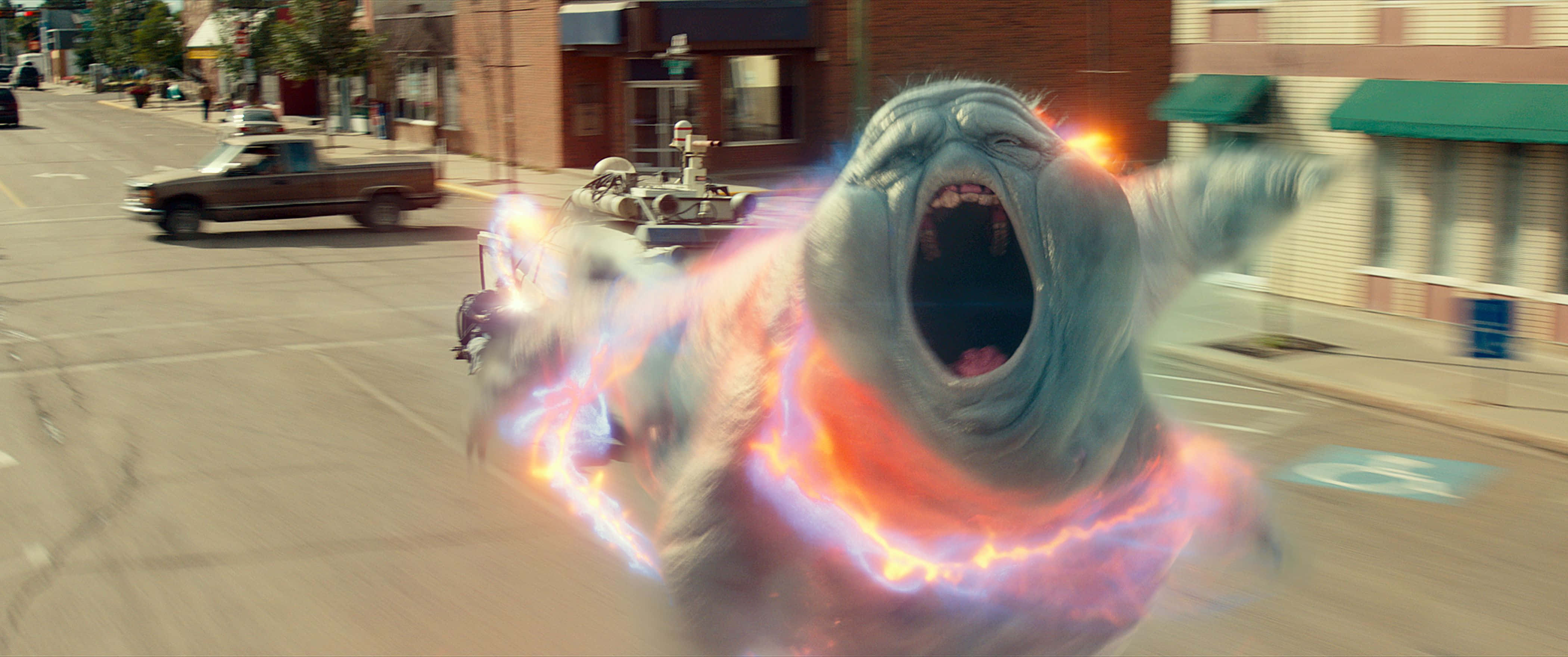 Ghostbusters 桌布 壁紙 — 一個巨大的幽靈生物，帶有發光的能量環，在城市的街道上高速奔馳並咆哮。