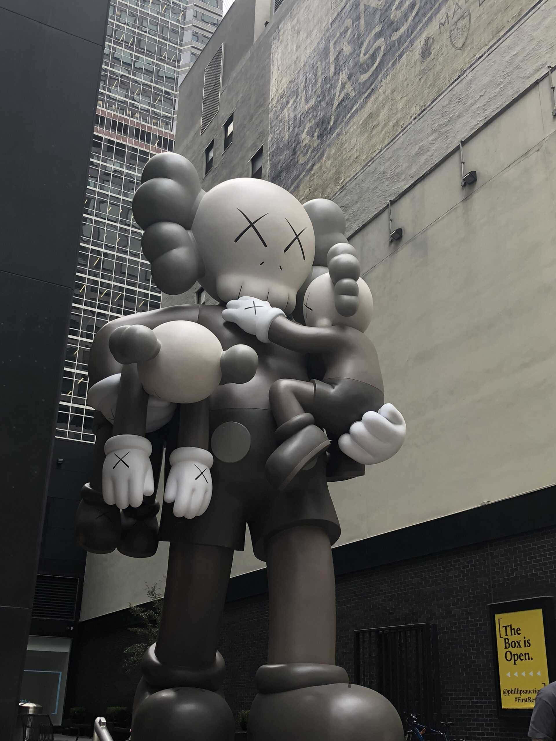 Kaws Holiday 桌布 壁紙 — 一個巨大的灰色KAWS雕塑，帶有X形眼睛，在高樓之間的都市小巷中擁抱著一個較小的版本。