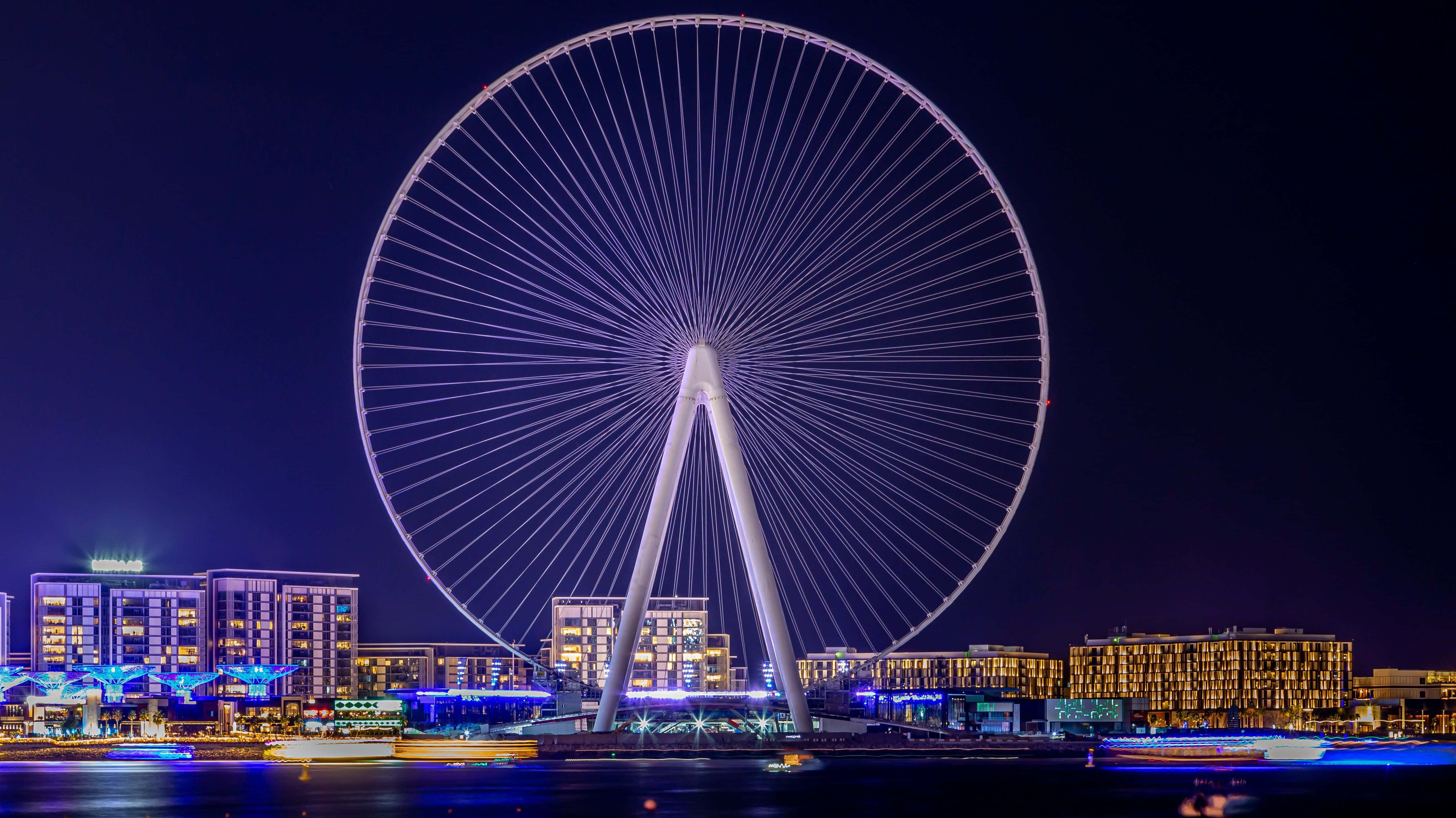 Uae 桌布 壁紙 — 一個巨大的燈光摩天輪主導著夜晚的海濱城市景觀，下方有發光的建築物和移動的船隻。