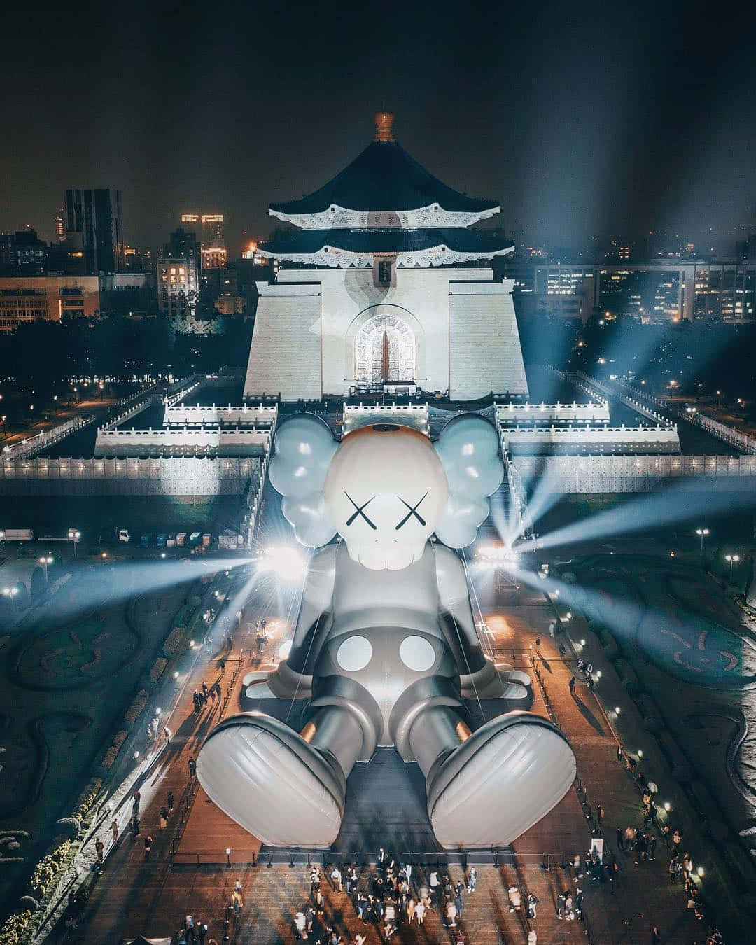 Kaws Holiday 桌布 壁紙 — 一個巨大的發光KAWS雕塑在夜間坐落於一棟燈火通明的傳統建築前，光束向外輻射。