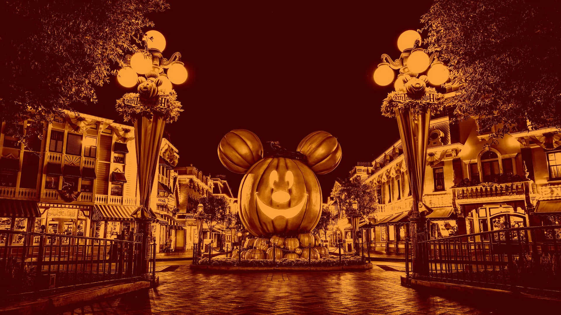 Disney Halloween 桌布 壁紙 — 一個巨大的發光南瓜燈，帶有米奇老鼠耳朵，在夜晚以溫暖的棕褐色調照亮節日氣氛的萬聖節街道。