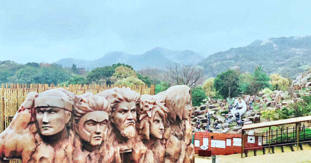 Naruto Village 桌布 壁紙 — 一個巨大的石雕，刻有五個表情強烈的戰士面孔，主宰著一個山丘公園，遠處有山脈和綠樹，天空朦朧。