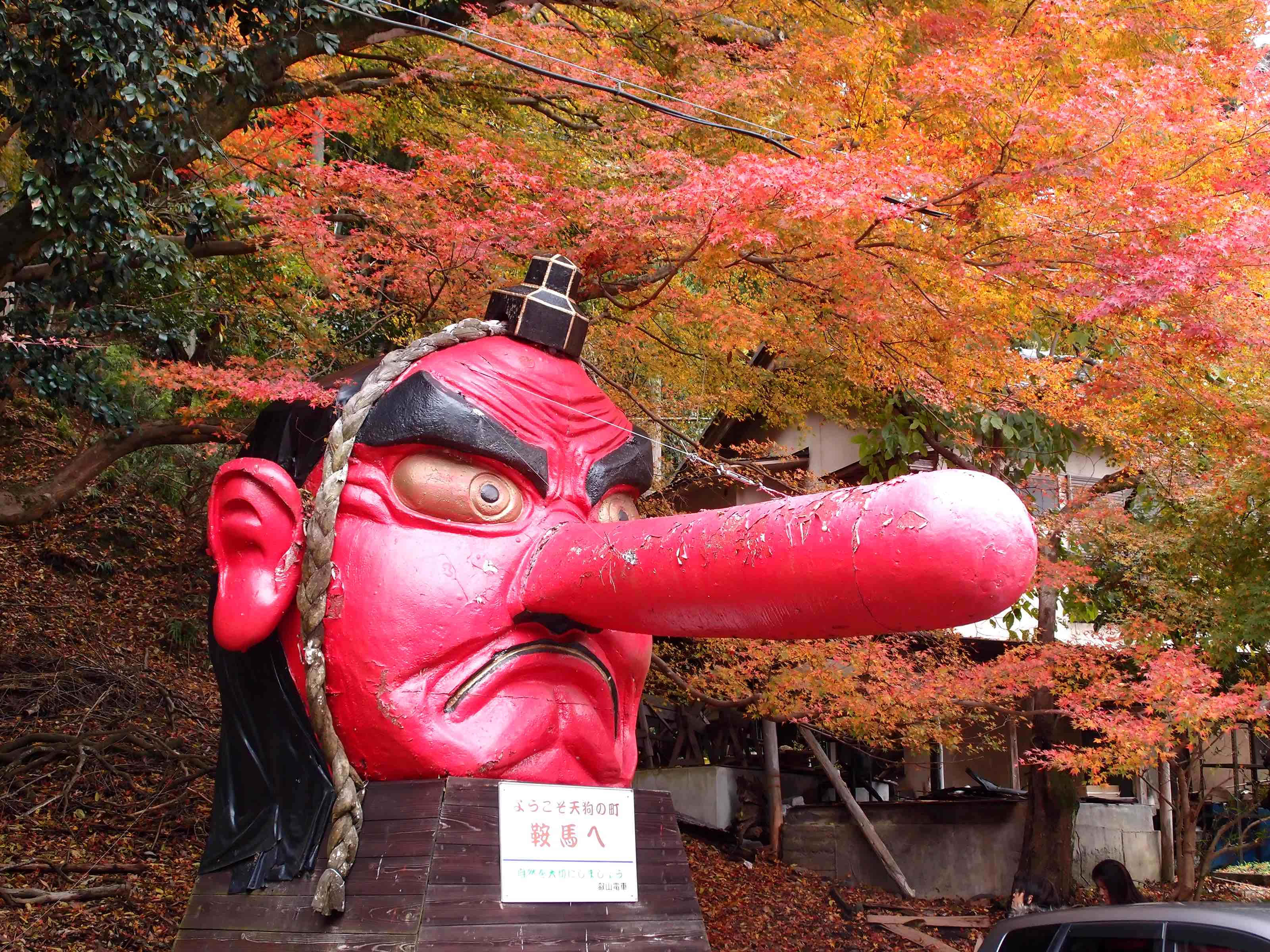 Tengu 桌布 壁紙 — 一個巨大的紅色日本鬼頭雕像，長鼻子，在日光下矗立在充滿活力的秋葉中。