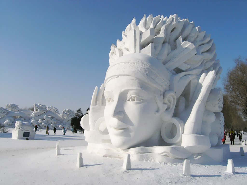 Ice Sculpture 桌布 壁紙 — 一個巨大而複雜的雪雕，雕刻著一張平靜的人臉，戴著精緻的頭飾，在晴朗的藍天下主導著明亮的冬季景觀。