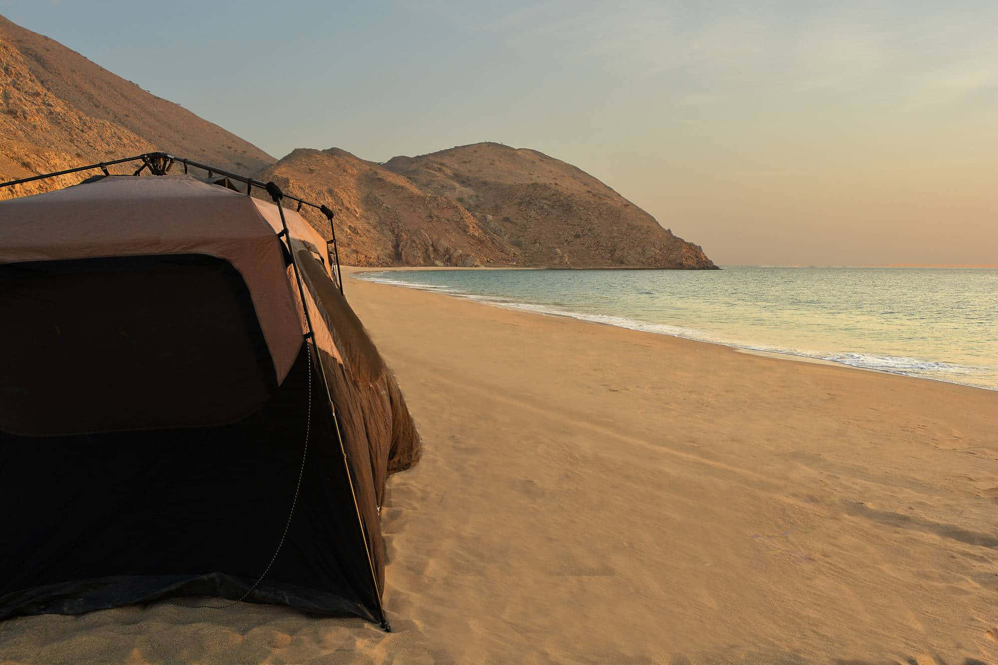 Beach Camping 桌布 壁紙 — 一個帳篷在日落時分搭建在沙灘上，背景是平靜的海浪和崎嶇的山丘。