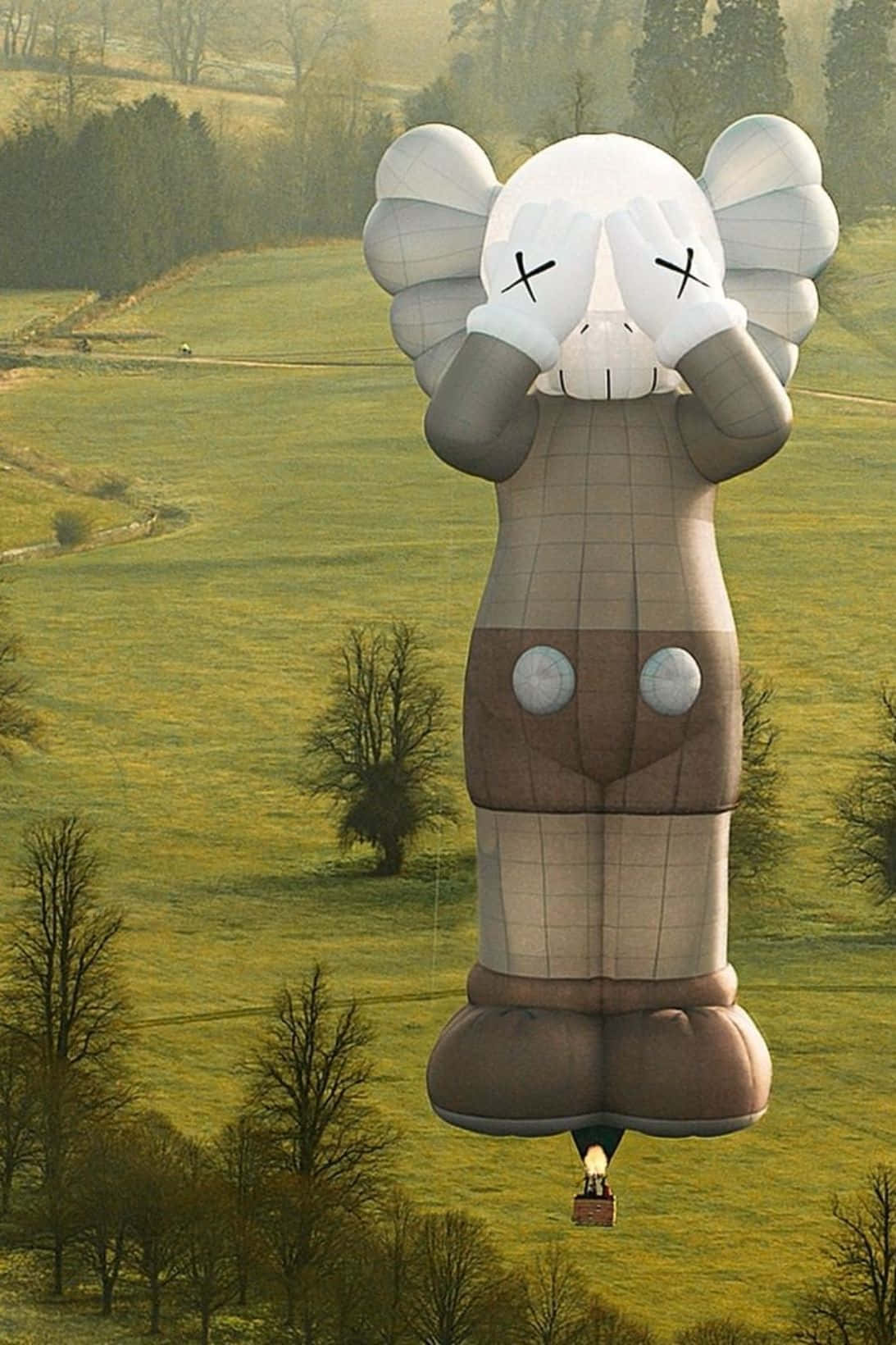 Kaws Holiday 桌布 壁紙 — 一個帶有X形眼睛的巨型KAWS氣球漂浮在陽光明媚、點綴著光禿樹木的綠色山谷上空。