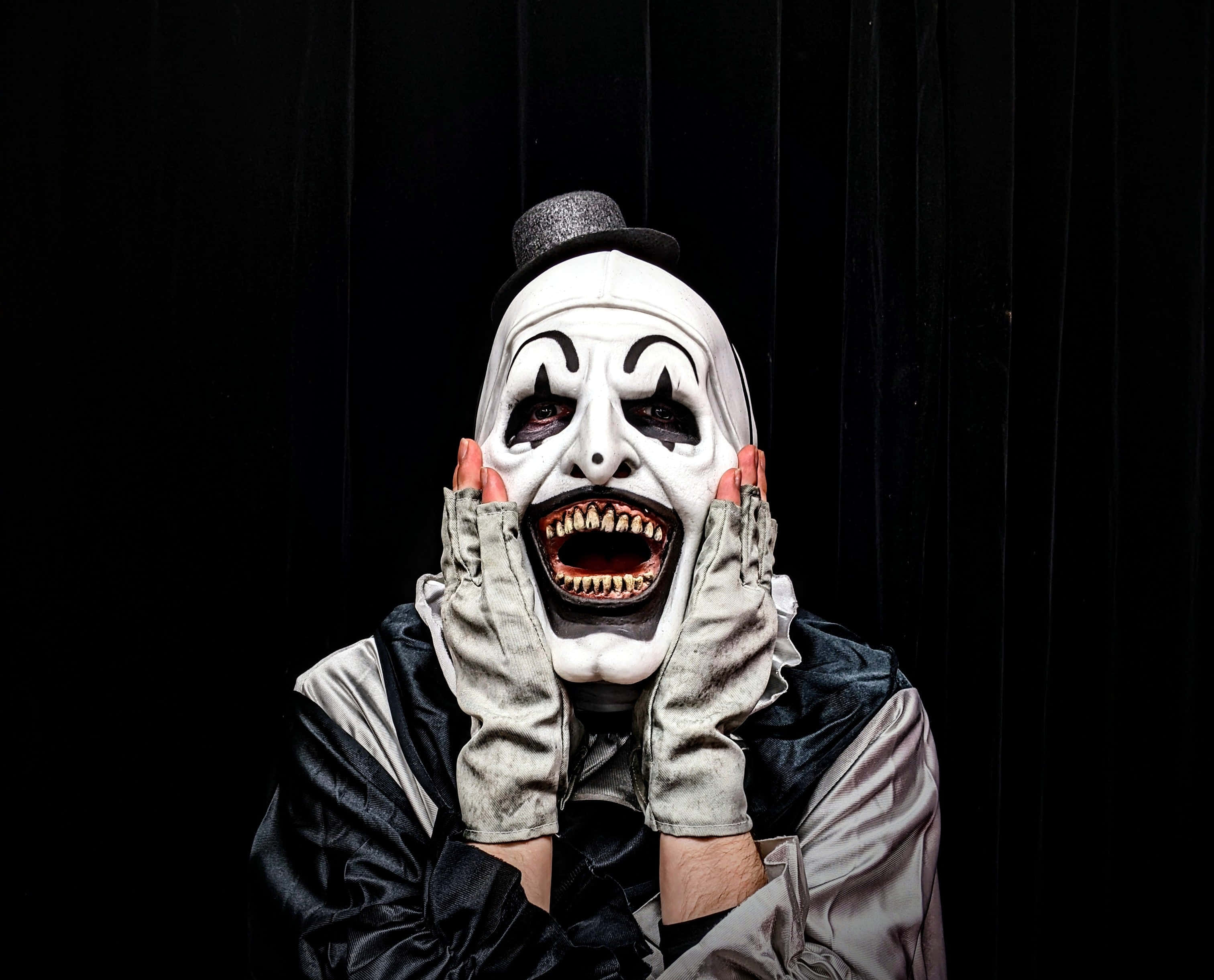 Halloween Masks 桌布 壁紙 — 一個帶有寬闊露齒笑容和紅色指甲的威脅性小丑，在黑色窗簾背景下將手按在臉上。