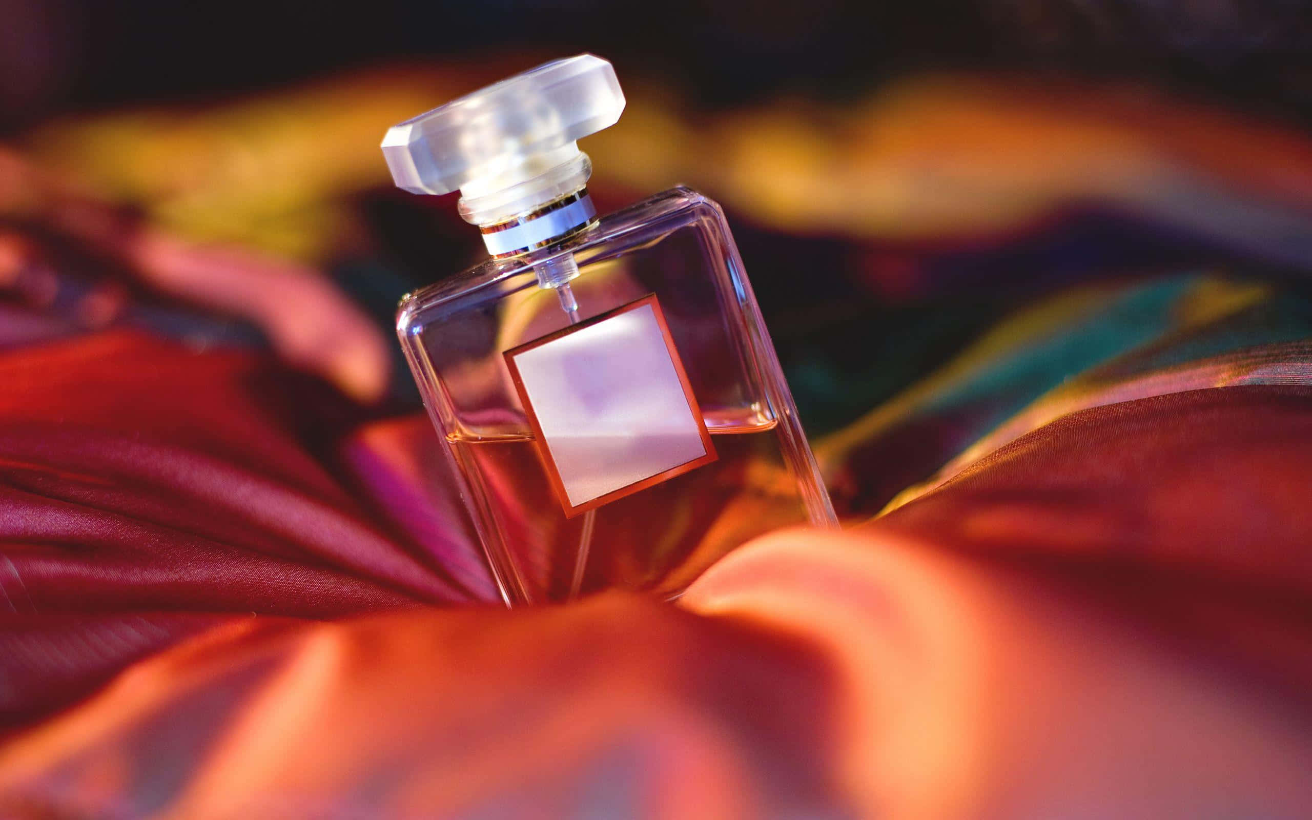 Floral Fragrance 桌布 壁紙 — 一個帶有幾何形瓶塞的透明香水瓶，優雅地放置在色彩豐富的緞面布料上，沐浴在溫暖的環境光中。