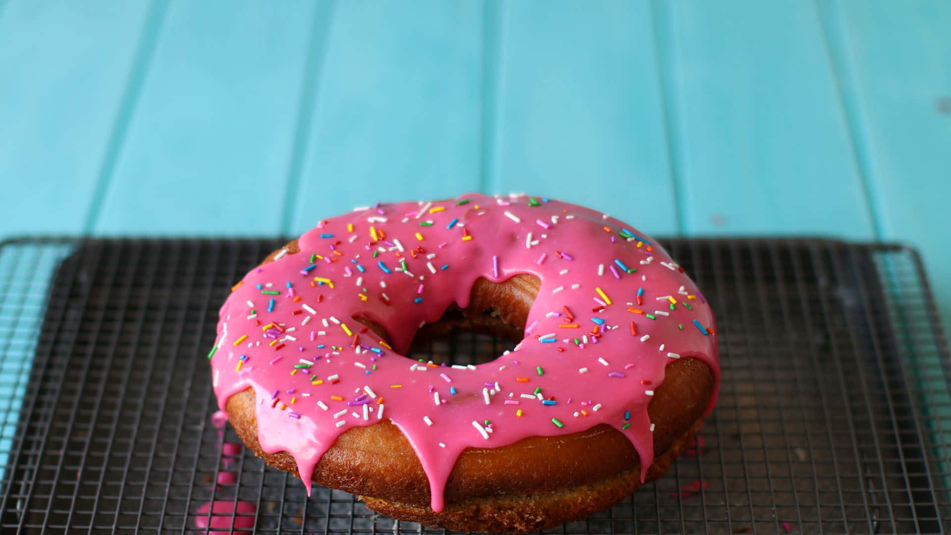 donuts 桌布 壁紙 — 一個帶有彩色糖粒的亮面粉紅糖霜甜甜圈，放在黑色冷卻架上，背景是明亮的藍綠色木質背景。