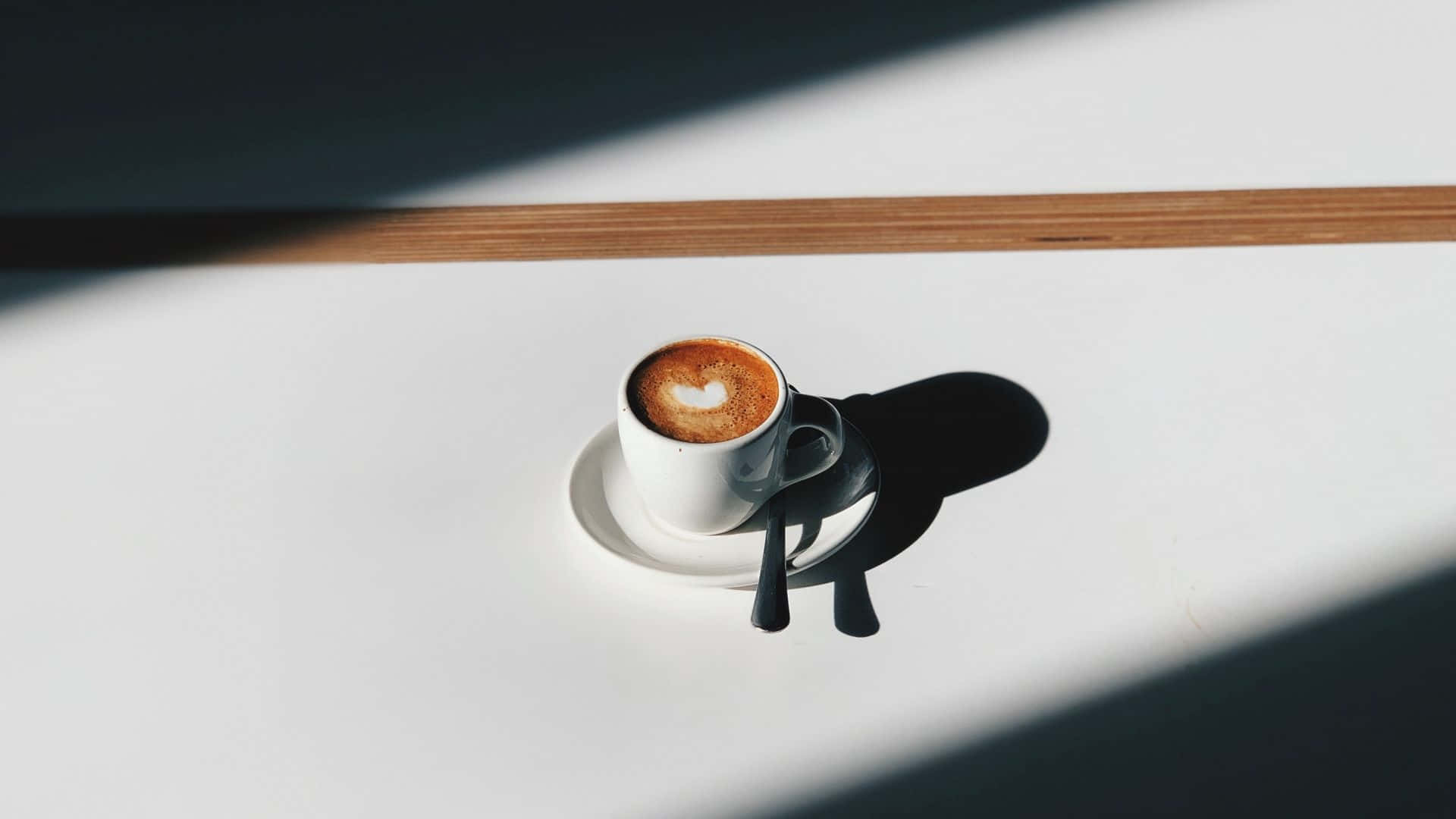 Minimalist Coffee 桌布 壁紙 — 一個帶有心形拉花藝術的白色咖啡杯放在碟子上，在強烈的陽光下於明亮的白色桌子上投射出清晰的陰影。
