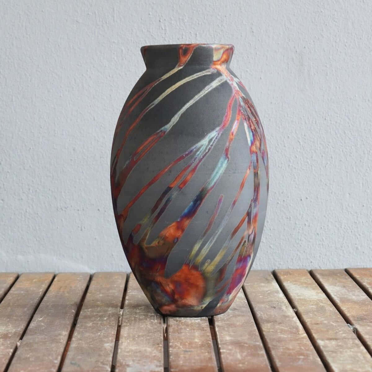 Ceramic 桌布 壁紙 — 一個帶有旋轉紅橙和藍色釉彩的陶瓷花瓶，放在一張風化的木桌上，背景是純白色的牆壁。