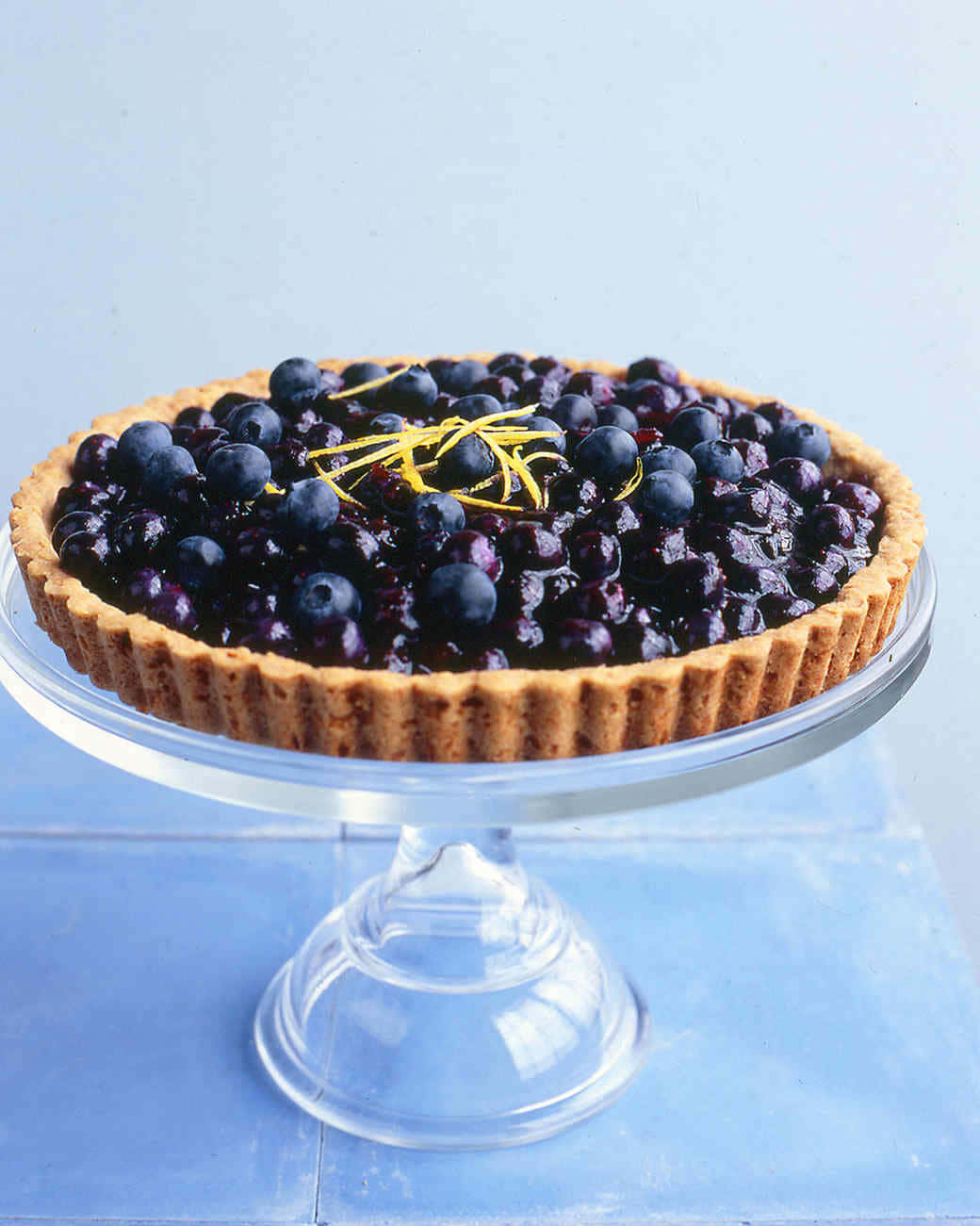 Blueberries Tart 桌布 壁紙 — 一個帶有檸檬皮的亮面藍莓撻放在玻璃底座上，背景是柔和的藍色。