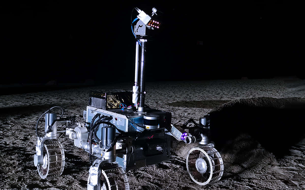 Lunar Rover 桌布 壁紙 — 一個帶有發光燈光的機械探測車站在黑暗、佈滿隕石坑的月球表面，天空漆黑一片。