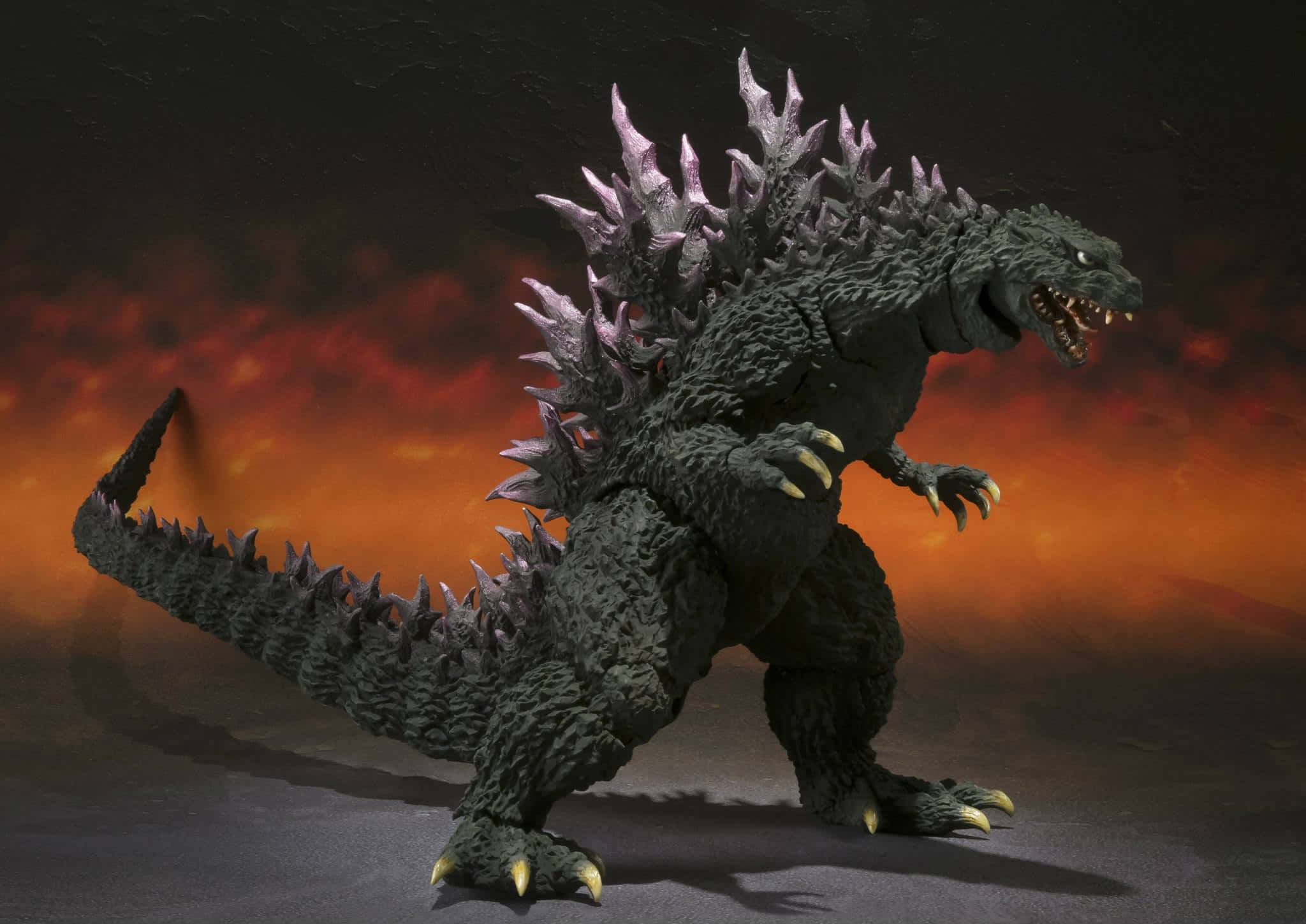 Godzilla 2000 桌布 壁紙 — 一個帶有發光眼睛和尖刺背部的威脅性哥斯拉人偶，站在熾熱的橙色和黑色天空前。