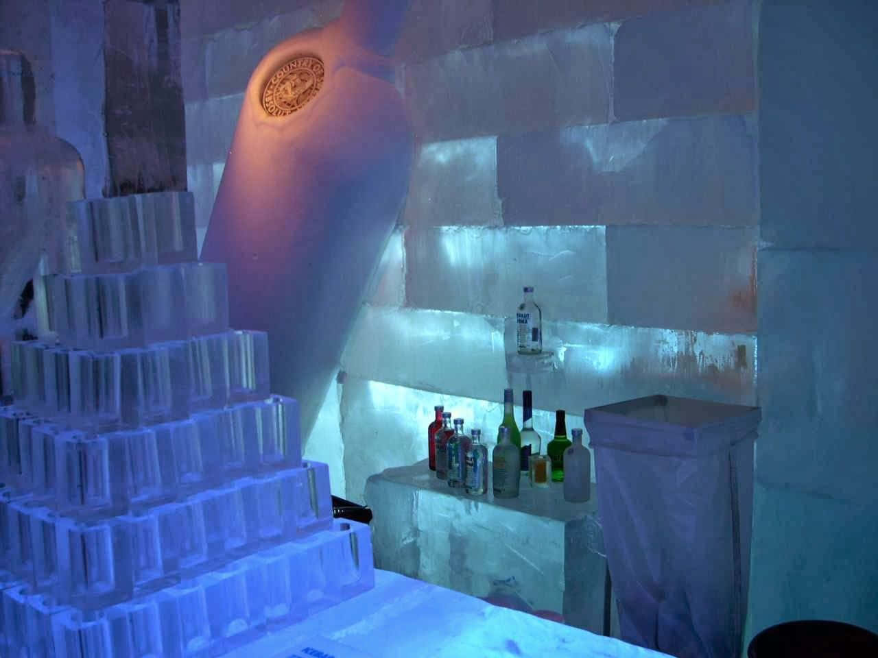 Ice Hotel 桌布 壁紙 — 一個帶有發光藍色燈光的冰吧，展示了一個巨大的冰瓶和冰杯金字塔，旁邊是一個擺放著酒瓶的吧檯。
