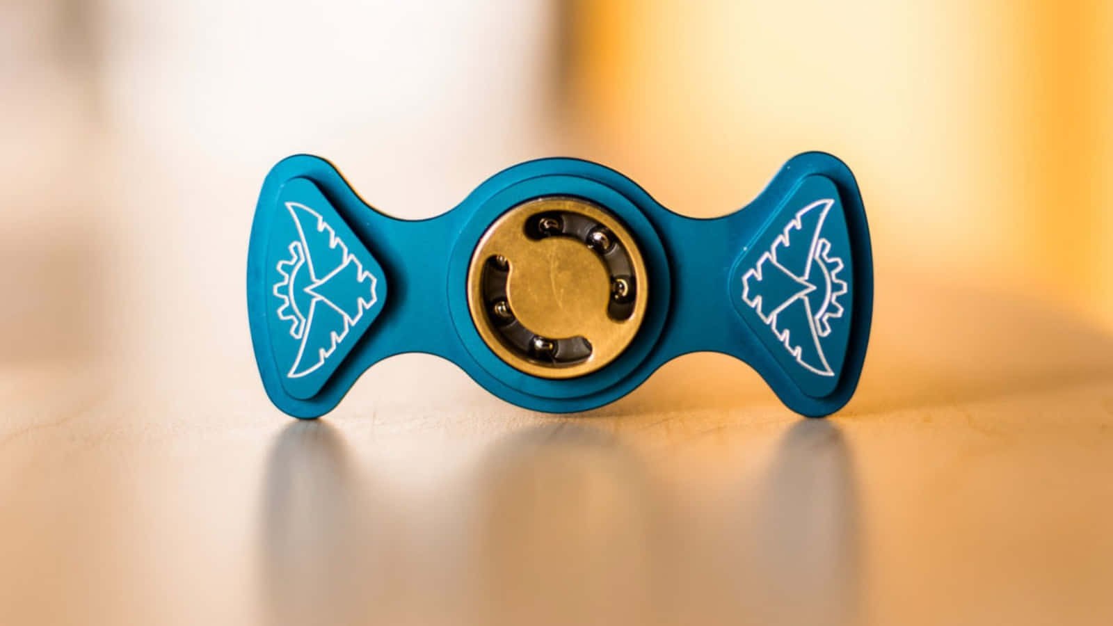 Fidget 桌布 壁紙 — 一個帶有白色部落圖案的青綠色指尖陀螺，在柔和的金色燈光下，放置在溫暖的木質表面上。