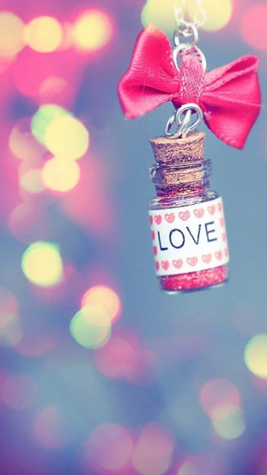 可愛少女風gif 桌布 壁紙 — 一個帶有粉紅色蝴蝶結、標有 LOVE 的小閃亮瓶子懸掛在色彩繽紛的散景背景前。